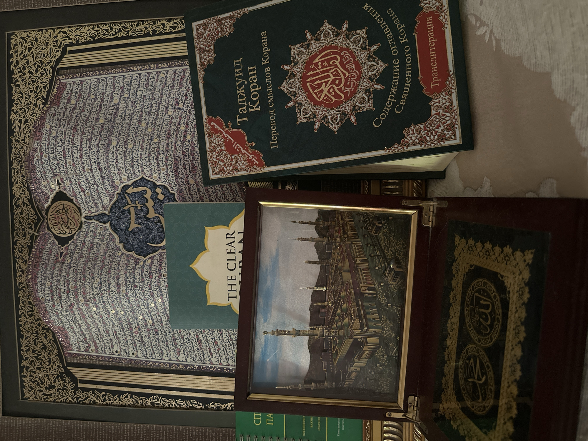 The image displays a collection of Islamic religious items. In the foreground, a wooden box with an ornate lid is open, revealing a decorative panel with Arabic calligraphy. To the right, a dark green book with gold lettering and intricate designs sits open, its title visible as "Таджуид Коран" (Tajweed Quran) and "Перевод смыслов Корана" (Translation of the Meanings of the Quran). A framed picture of a mosque, likely the Prophet's Mosque in Medina, is positioned in front of other books. In the background, a large framed artwork features extensive Arabic calligraphy. The overall impression is of a display of religious texts and art, possibly in a private home or a religious institution. The image does not contain any recognizable landmarks or elements that would definitively place it in Dushanbe, Tajikistan.