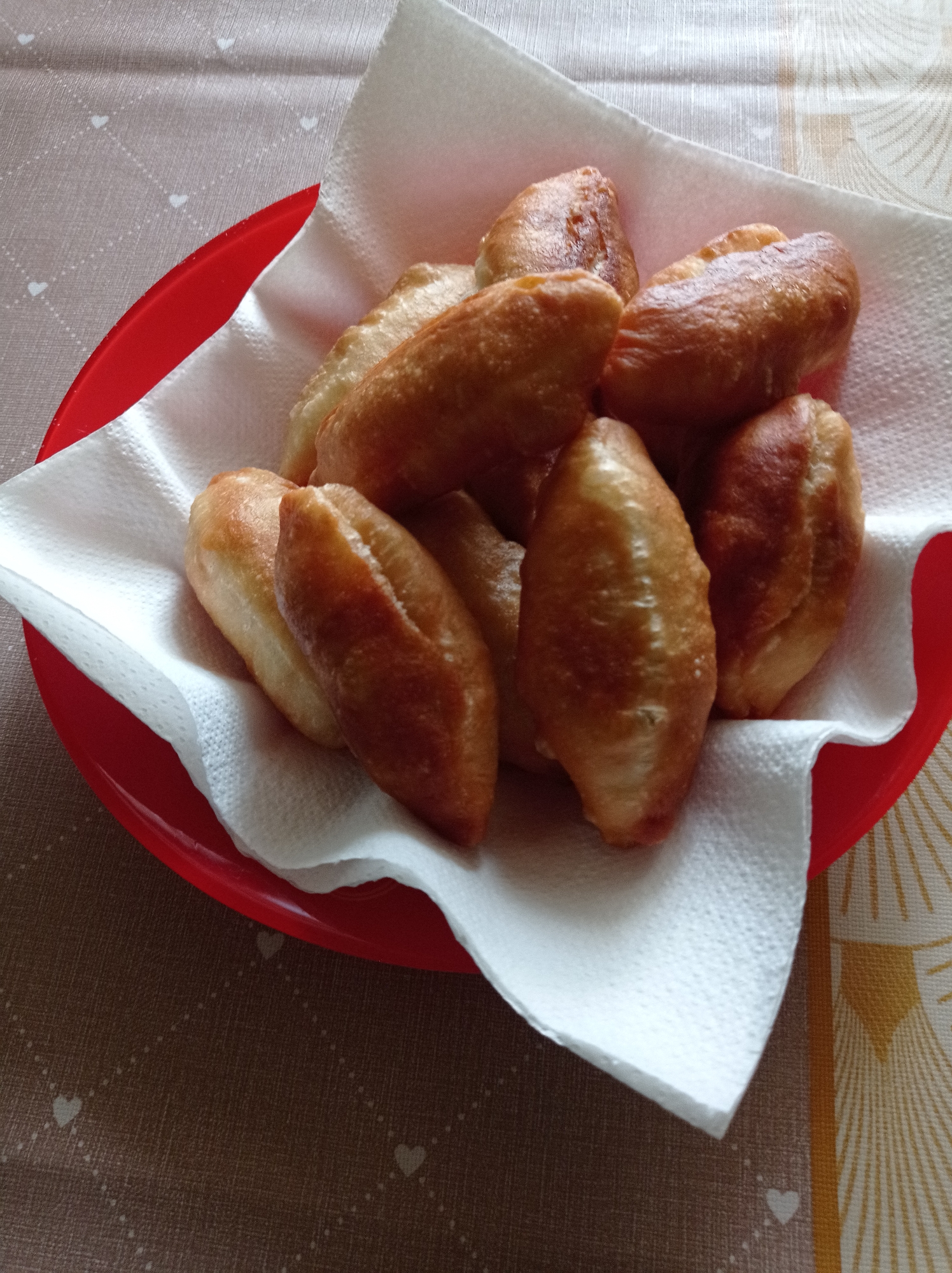 A close-up shot shows a red plate lined with a white paper towel, containing approximately nine fried pastries. The pastries are golden brown, slightly elongated, and appear freshly cooked. The plate is placed on a tablecloth featuring two distinct patterns: one side has a brown background with small white heart shapes, while the other side has a beige background with a stylized sunburst or fan pattern in shades of gold and brown. The location of Degtyarsk, Russia, is not discernible from the provided image.