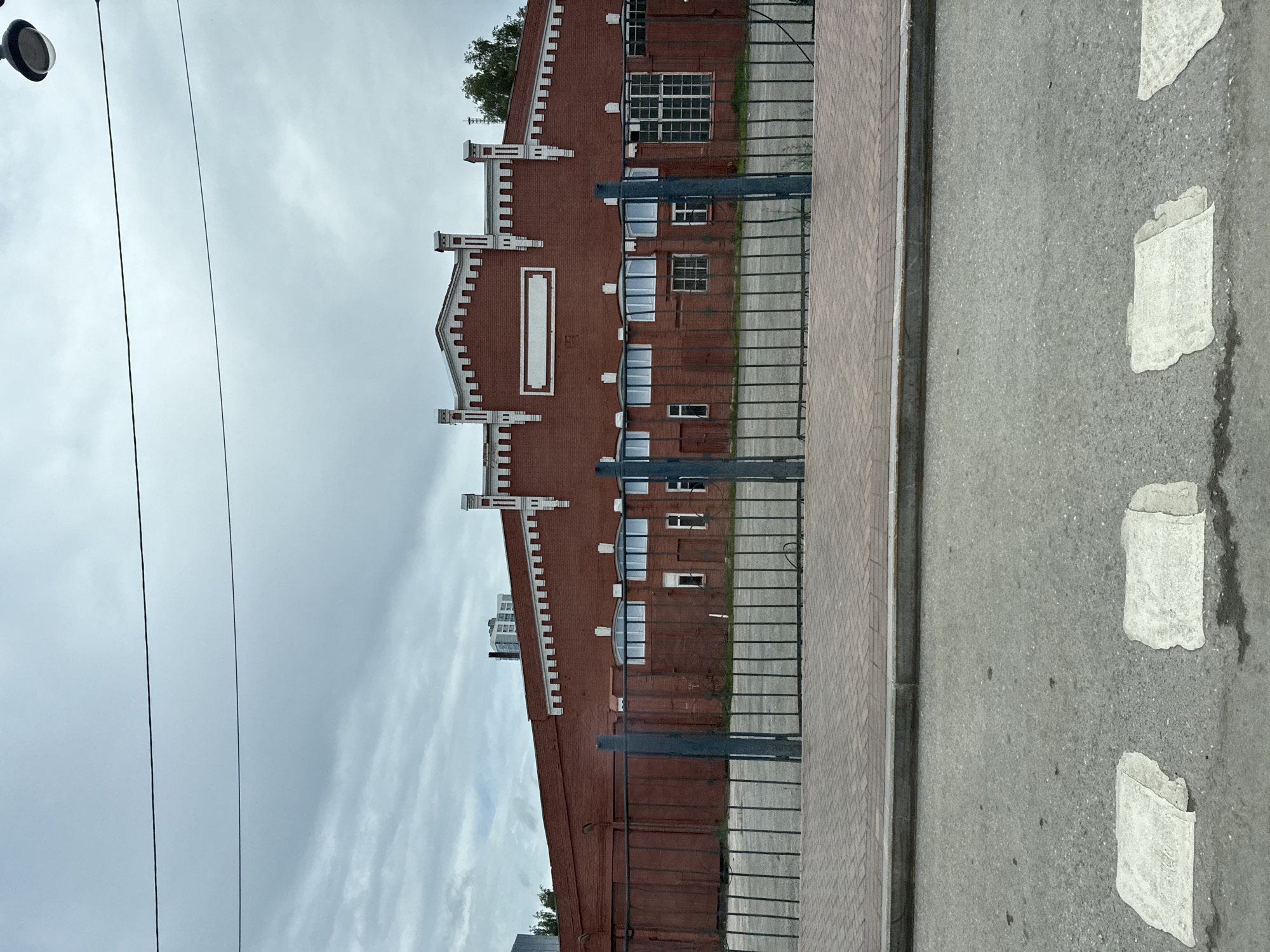 The image captures a wide shot of a long, red brick industrial building with a decorative white facade under an overcast sky. A dark metal fence with vertical bars separates the foreground from the building. The fence is set back from a paved sidewalk, which is in turn bordered by a street with a pedestrian crossing marked by two faded white stripes. The building features numerous windows and large, dark doors. A few trees are visible behind the fence on the right side of the building. The scene appears to be in an urban environment, likely a commercial or industrial area. The overall atmosphere is somewhat somber due to the cloudy weather.