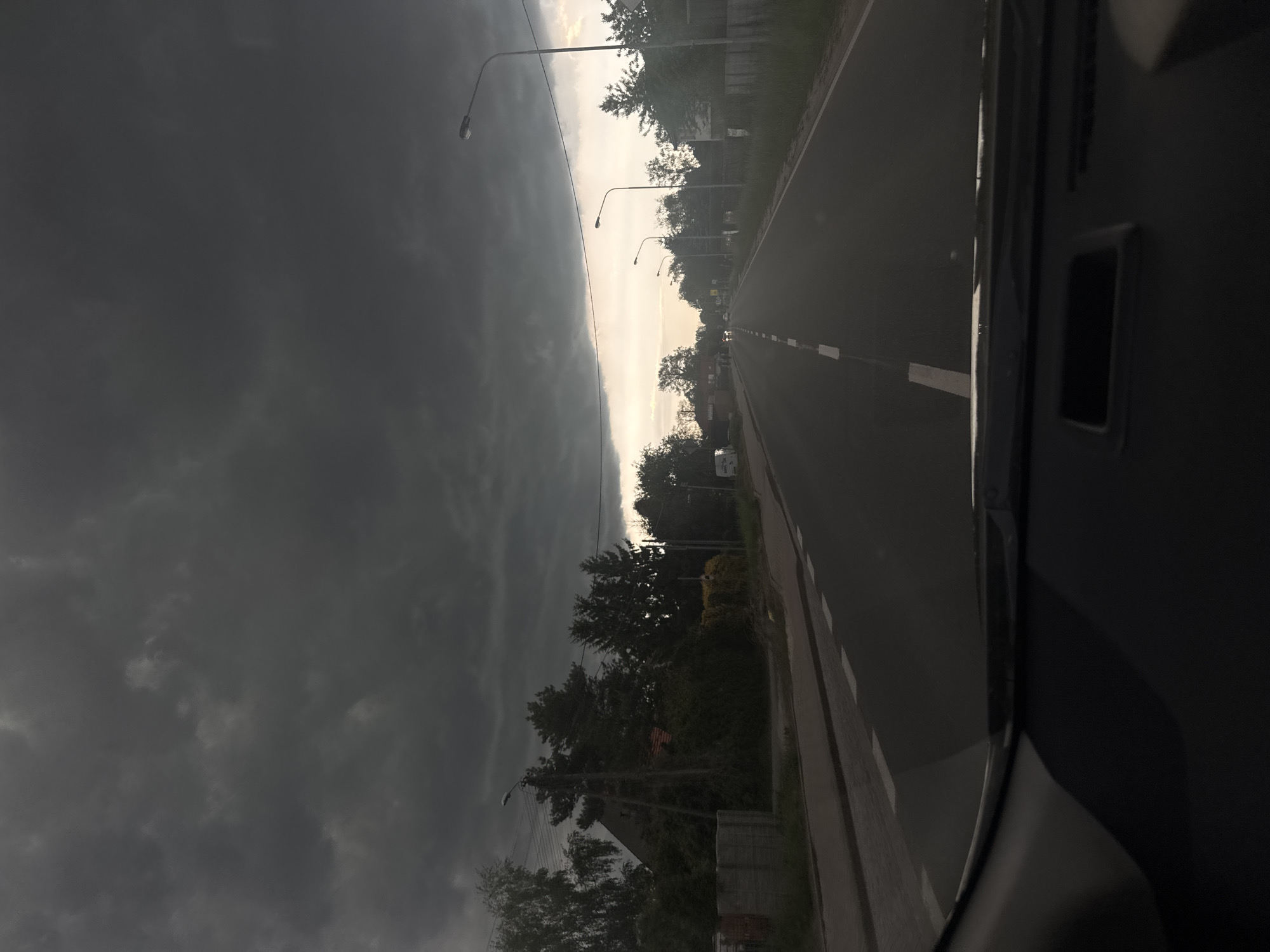 The image depicts a road in an unknown city, observed from inside a vehicle. The road is straight and has a white centerline. Street lamps line the road at regular intervals. Buildings and trees are visible alongside the road. The sky is split, with a dark, stormy cloud cover on the left and a lighter, clearer sky on the right. The weather appears unsettled, possibly with an approaching storm. There are no people visible in the scene.
