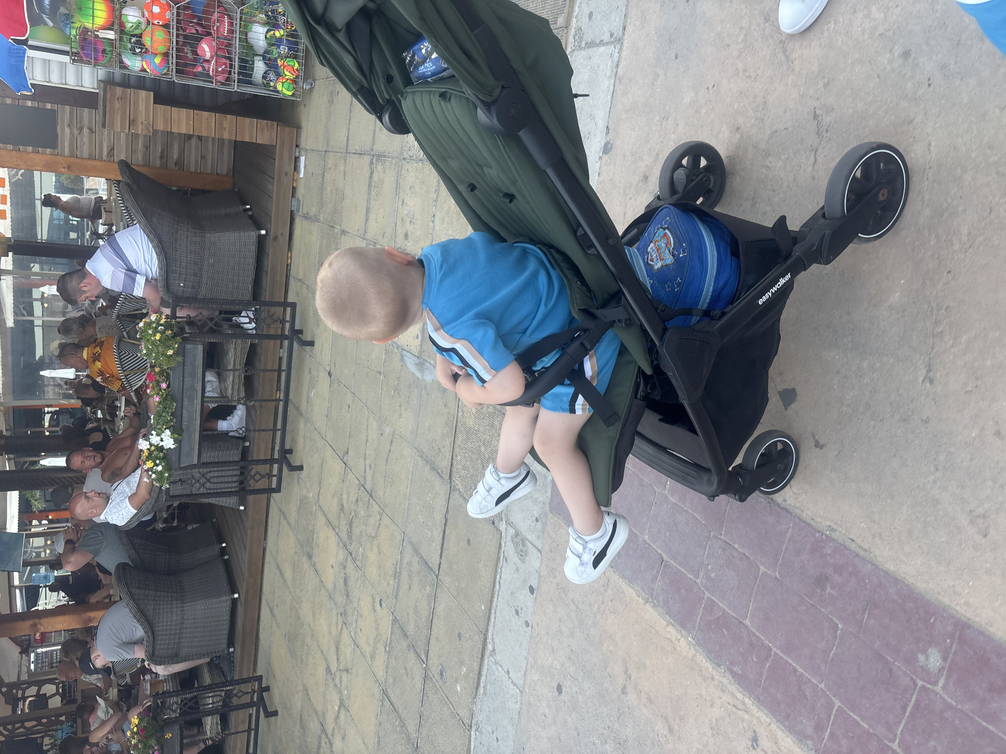 The image depicts a sunny outdoor scene in Nesebar, Bulgaria, likely at an establishment with outdoor seating. In the foreground, a young child with blond hair is seated in a dark green stroller, wearing a blue t-shirt with striped sleeves and white velcro sneakers. The stroller's branding, "easywalker," is visible on the frame. Beneath the stroller, a blue bag with a Paw Patrol design is visible.
In the background, several adults are seated at tables, some indoors and some outdoors. The seating area appears to be a patio or terrace with tiled flooring. To the right, a display rack holds colorful balls. The overall impression is one of a casual, public gathering place, possibly a restaurant or cafe, during daylight hours.
