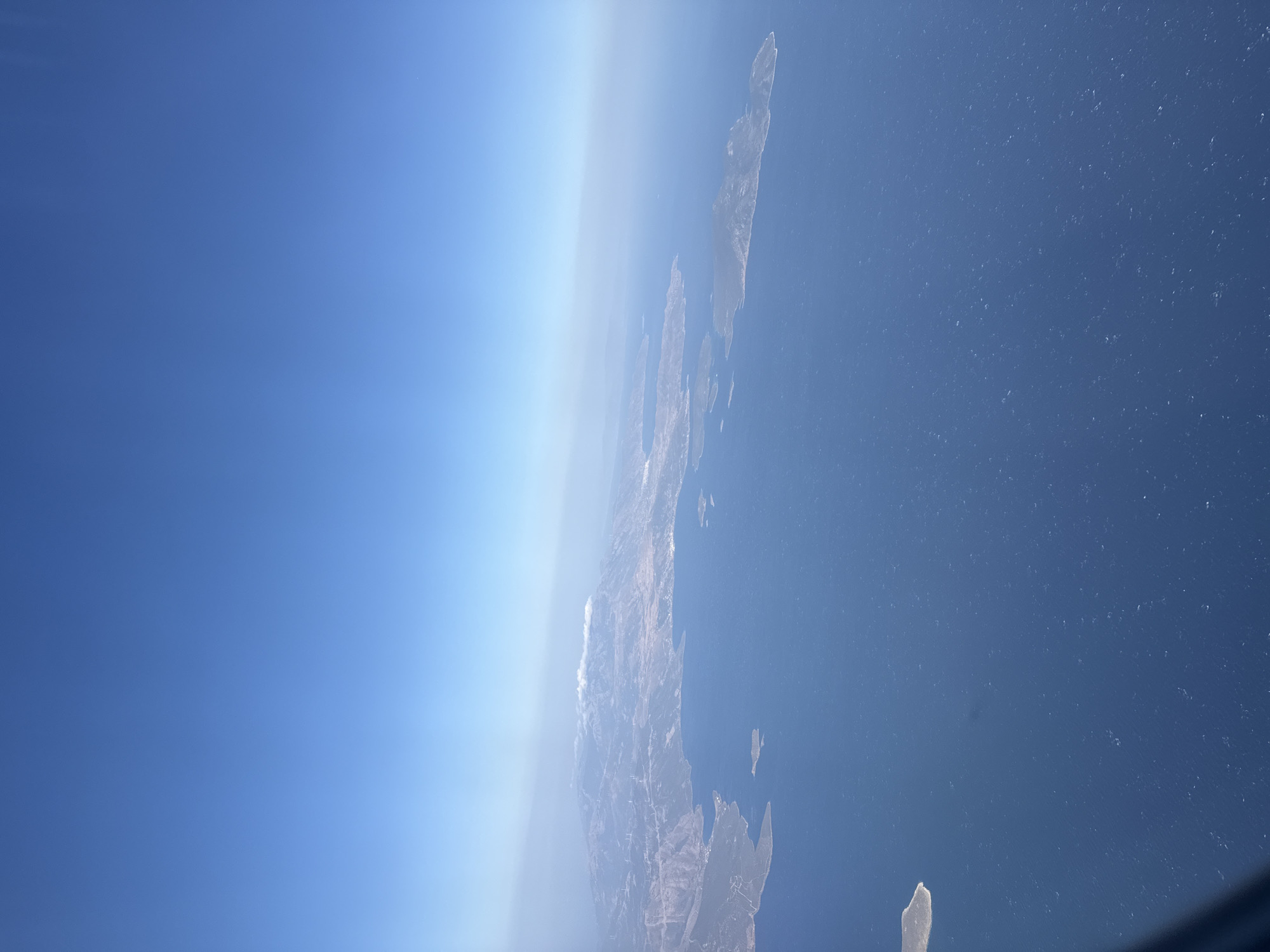 This is an aerial photograph showing a coastal region of Greece. The image features a vast expanse of deep blue ocean in the foreground, with several islands and a mountainous mainland visible in the distance. The mainland is characterized by rugged terrain, with some areas appearing arid and others covered in vegetation. Wind turbines are scattered across the hills of the mainland, indicating a source of renewable energy. A plume of white smoke rises from the crest of one of the mountains, possibly indicating a wildfire or industrial activity. The sky is a clear, bright blue, with a slight haze visible near the horizon. The overall impression is one of a natural landscape meeting human civilization. The specific location is unknown, but the scenery is typical of the Aegean islands or coastal regions of mainland Greece.
