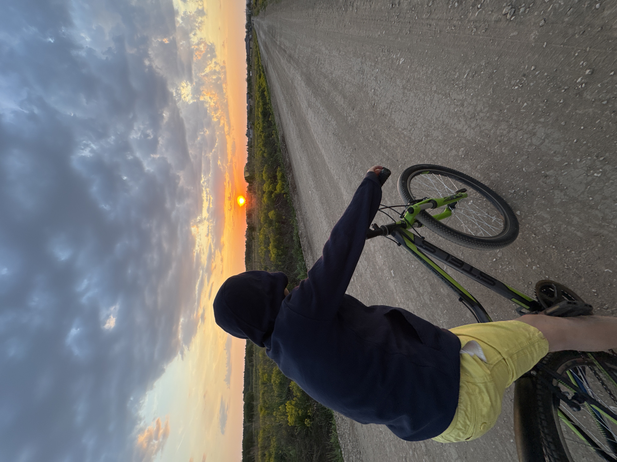 A low-angle, wide shot captures a person riding a bicycle on a dirt road at sunset in Ферма, Russia. The individual, facing the setting sun, wears a dark blue hooded sweatshirt and yellow shorts. The bicycle is mostly black with green accents. The road stretches towards the horizon, flanked by vegetation on either side. Above, a dramatic sky features a mix of grey clouds and orange hues from the setting sun. The overall scene conveys a sense of solitude and rural tranquility.
