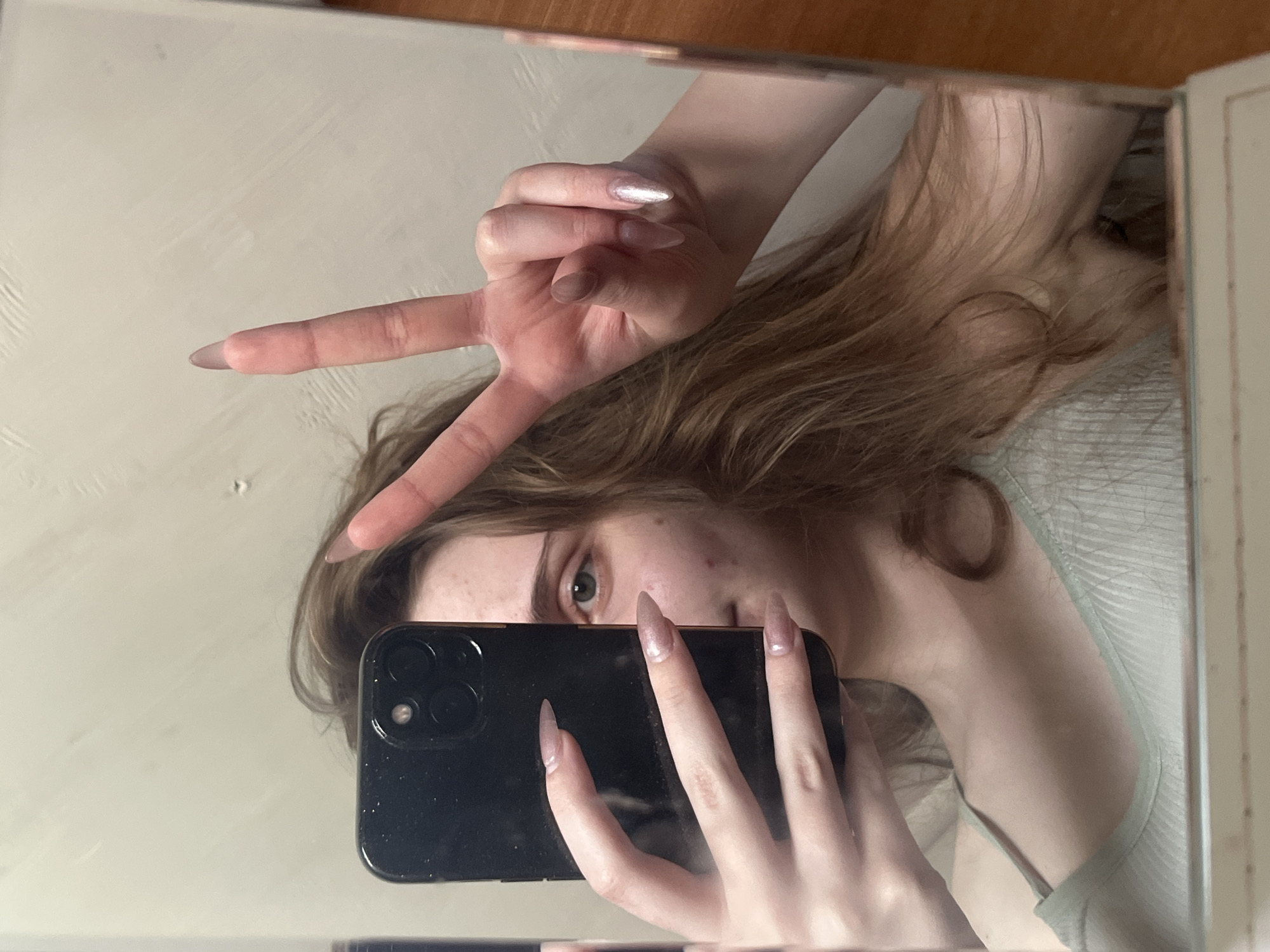 In a reflection, a young woman with long, wavy brown hair is holding a black smartphone in her left hand, partially obscuring her face. Her right hand is raised, forming a peace sign. She appears to be wearing a light green ribbed tank top. The background is a plain, light-colored wall. The scene is indoors, likely a bathroom or bedroom, and there are no discernible elements that place it specifically in Ashkadar, Russia.