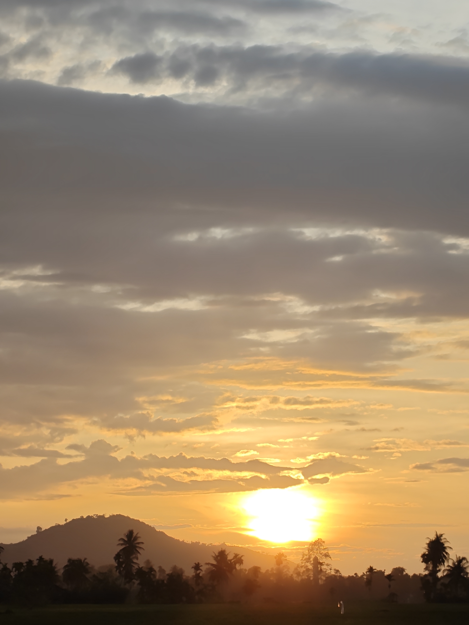 The image depicts a sunset over a landscape in Meunasahalue, Indonesia. The sun is positioned near the horizon, partially obscured by clouds, with a bright golden glow illuminating the sky. Silhouettes of a hill and trees, including palm trees, are visible in the foreground. Darker clouds are present in the upper portion of the sky. A figure of a person is barely visible in the field.
