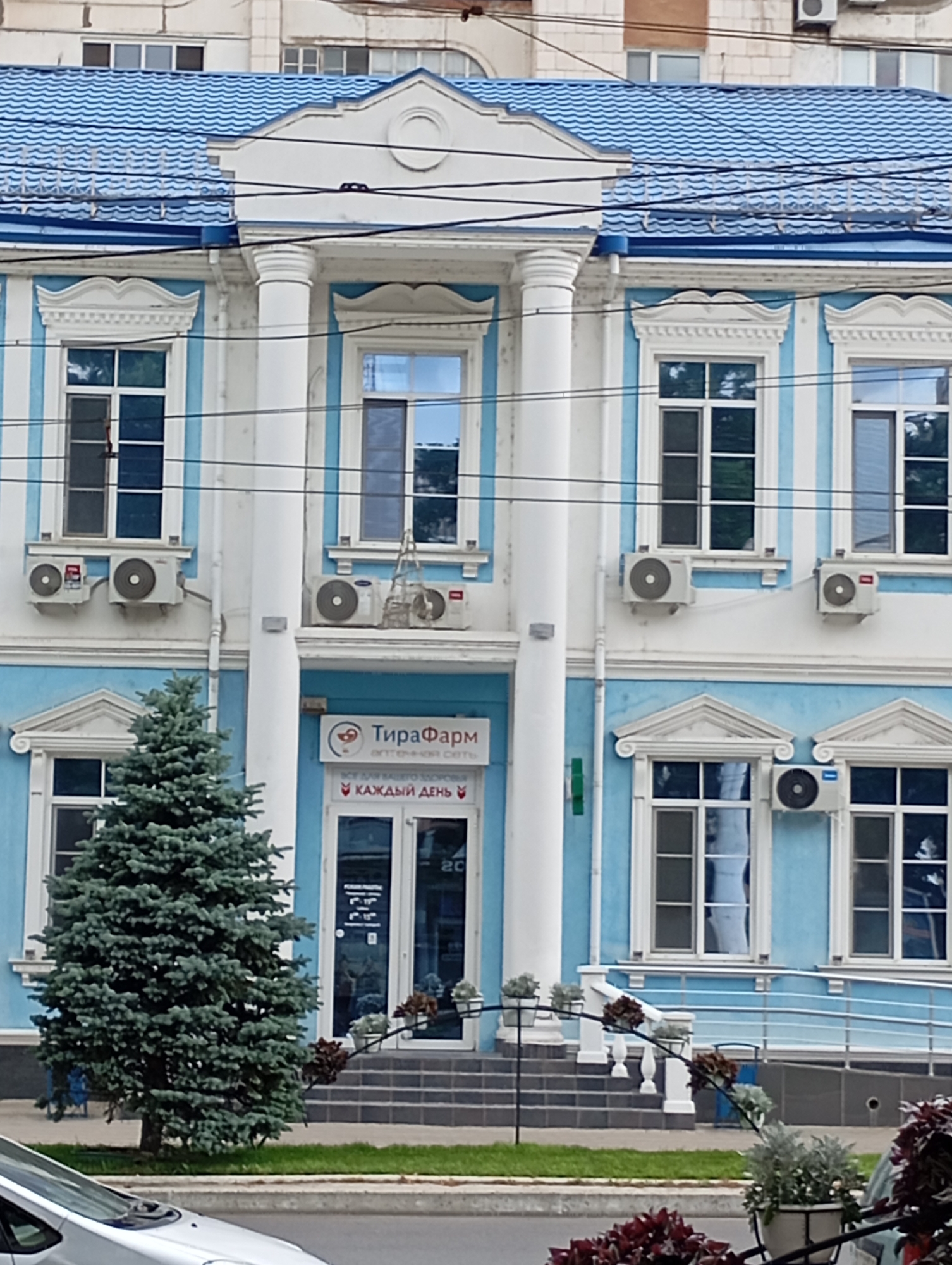 The image shows the facade of a light blue and white building in Tiraspol, Moldova. The building has a blue tiled roof and several large windows with white trim. There are air conditioning units mounted on the exterior walls below some of the windows. A small evergreen tree is planted in front of the building, to the left of the entrance. The entrance features double glass doors with a sign above that reads "ТираФарм" and below that "КАЖДЫЙ ДЕНЬ". A silver car is partially visible in the lower left corner of the frame. The scene appears to be on a street with a sidewalk and grass in front of the building.