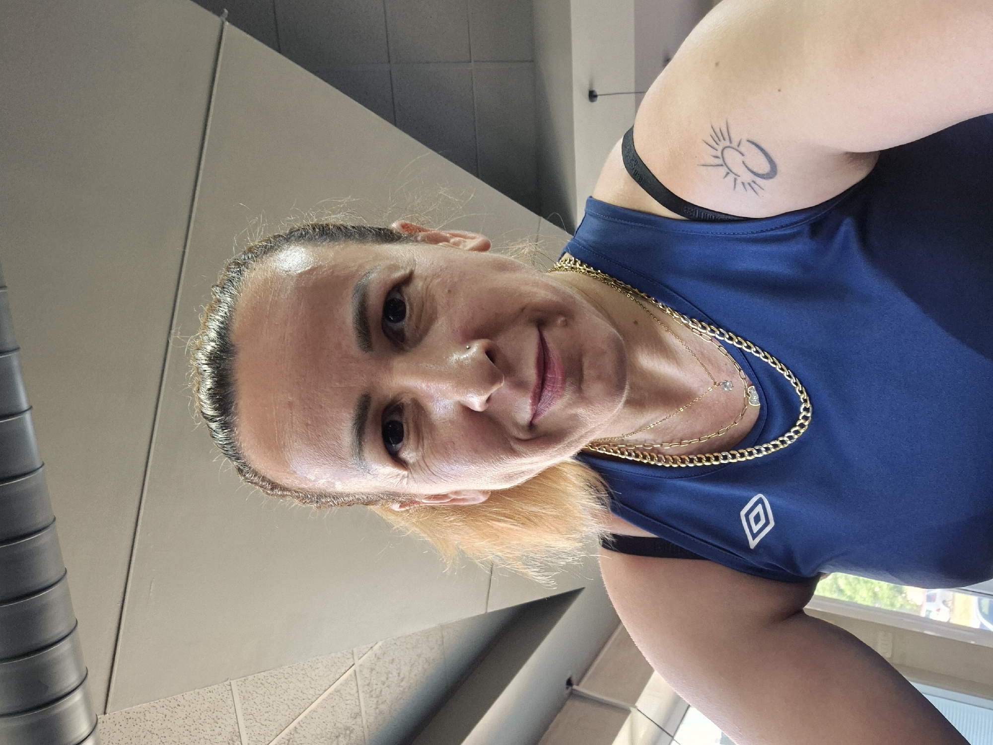 A woman with blonde hair pulled back and a tattoo of a sun and crescent moon on her upper right arm is captured in a close-up, selfie-style shot. She is wearing a dark blue athletic tank top with a white logo on the left chest and a black sports bra underneath. Two gold necklaces adorn her neck. The background consists of a grey ceiling with industrial ductwork and light-colored walls, suggesting an indoor setting like a gym or an industrial space. The lighting is bright, indicating daylight.