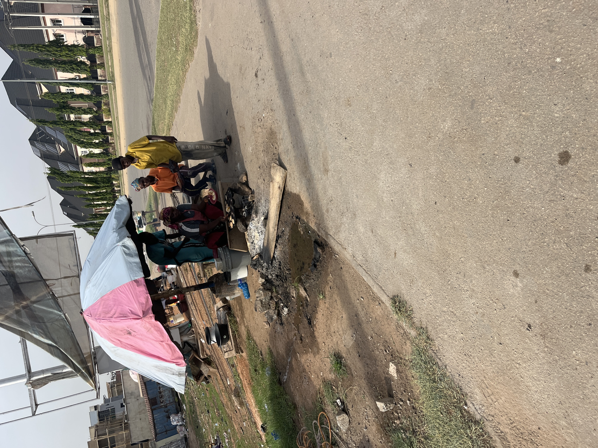 The image shows a roadside scene in Karum, Nigeria. Three men are gathered around a small cooking setup. One man is standing, wearing a yellow shirt and Old Navy pants. Two other men are seated. A fire is burning near them, and there are cooking pots and other equipment. A large, pink and white striped umbrella provides shade. The surroundings appear to be a mix of pavement, dirt, and sparse vegetation. In the background, houses are visible.