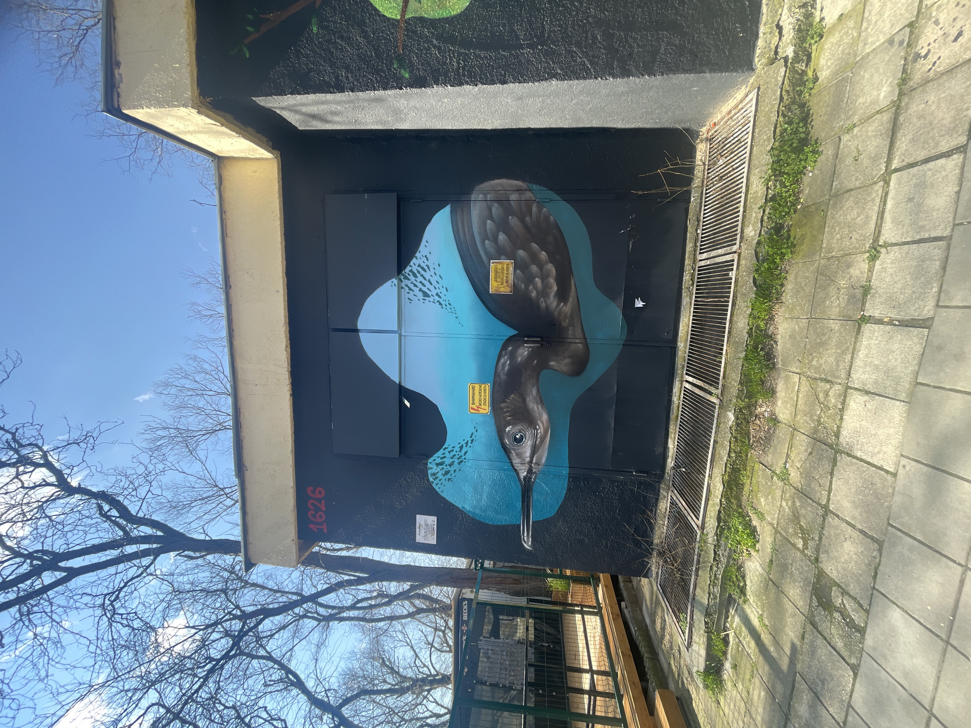 The image depicts a street art mural on a utility box in Varna, Bulgaria. The mural portrays a bird with grey and black plumage, set against a blue background resembling water. The bird’s head is oriented downwards. The box itself is painted black, with the number "1626" written in red on the side. Warning labels in Cyrillic script are affixed to the artwork. The utility box is located next to a sidewalk paved with concrete tiles, where grass and weeds are growing in the crevices. A metal grate is visible on the side of the box. Bare tree branches are visible in the background against a clear blue sky. A partial view of a metal fence and wooden beams suggests a nearby construction site or enclosed area.