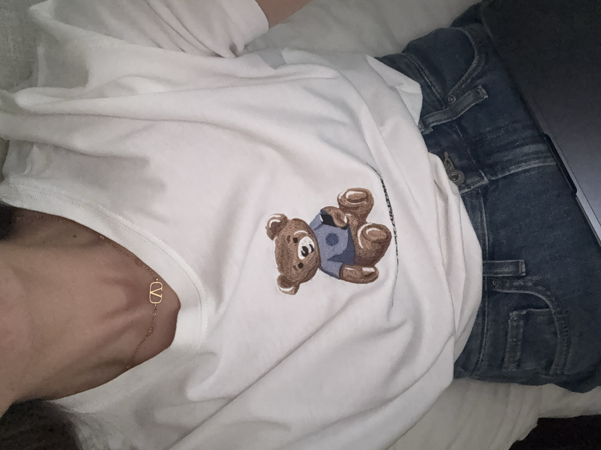 The image shows a person lying on a white surface, possibly a bed, wearing a white t-shirt with a cartoon teddy bear embroidered on it and blue jeans. A gold Valentino necklace is visible on the person's neck. A laptop is partially visible on the right side of the image, near the person's hip. The location is not discernible from the image content. Therefore, it is impossible to determine the location is the city of San Bernardino Tlaxcalancingo, Mexico, or anywhere else.