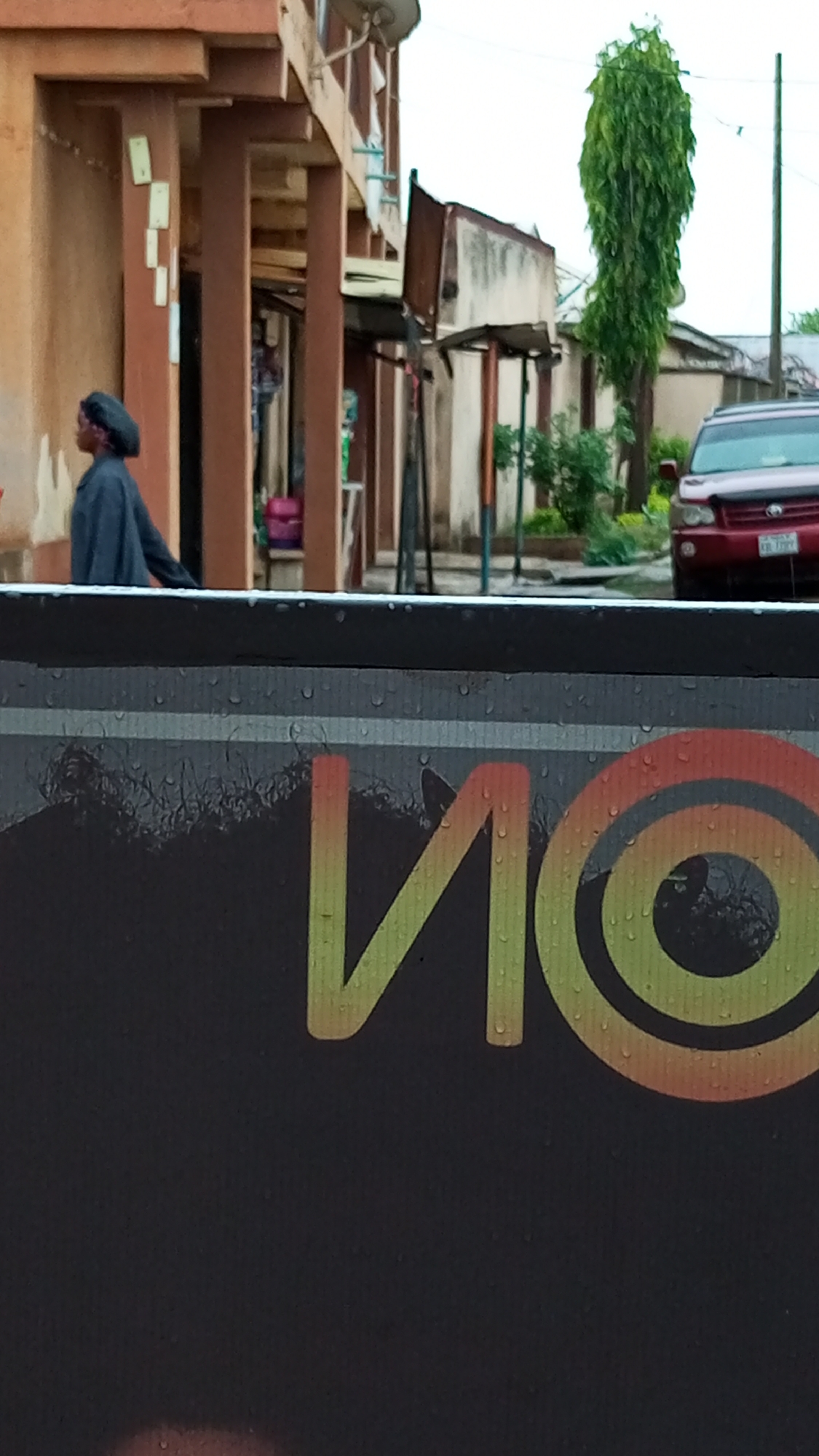 A street view in Sabon Tasha, Nigeria, shows a partially visible sign in the foreground with the word "NO" in yellow and red lettering. Behind the sign, a woman in a dark head covering and dark clothing walks along the side of a building with several pillars. Further down the street, a red SUV is parked, and a tall, slender tree is visible. The buildings are simple structures. Overhead, the sky is overcast, suggesting a wet or humid climate.
