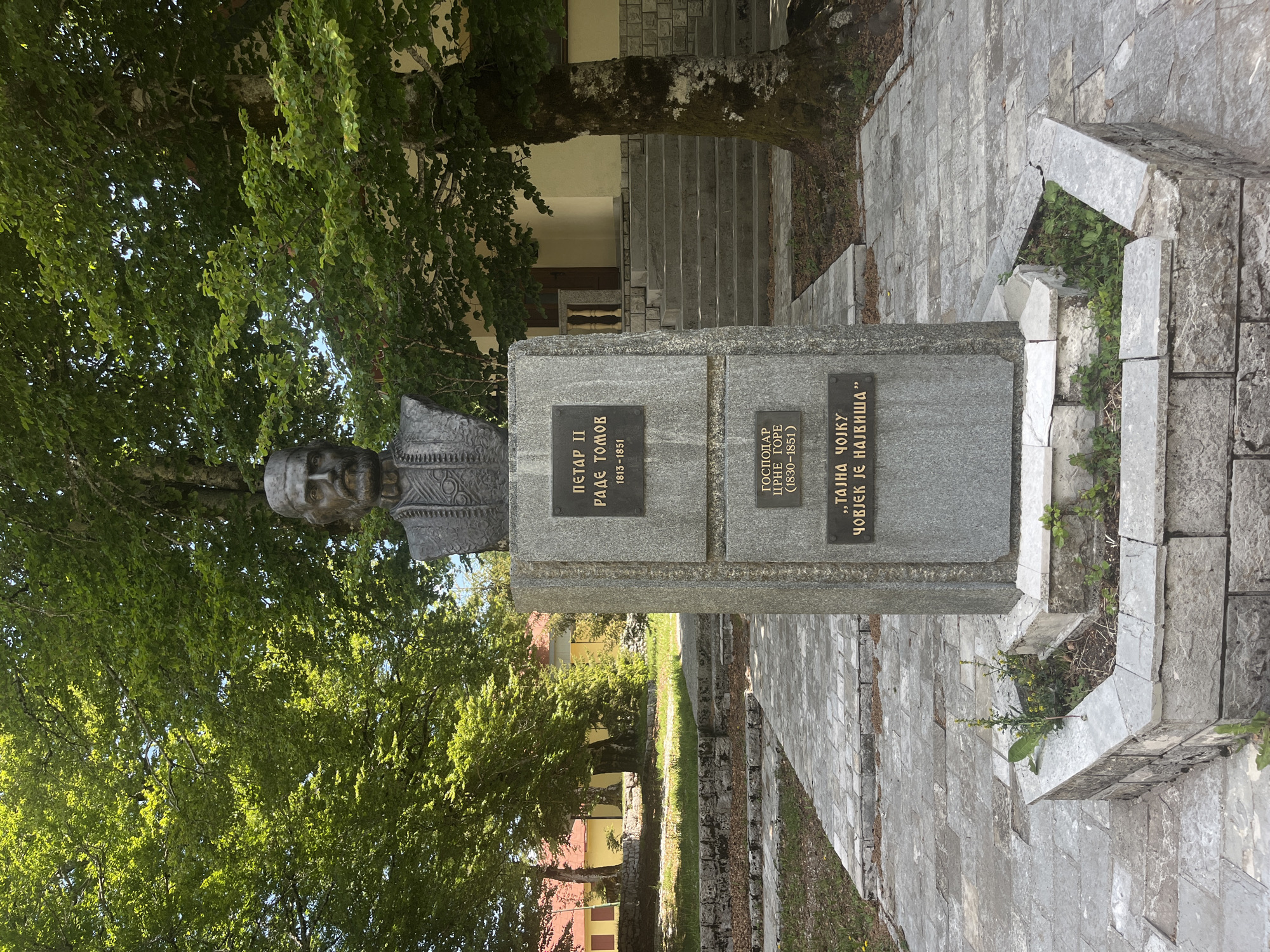 The image shows a stone bust erected in Velji Bostur, Montenegro. The bust is placed atop a stone plinth with two name plates, one reading "ПΕΤΑΡ ΙΙ РАДЕ ТОМОВ 1813-1851" and the other "ГОСПОДАР ЦРНЕ ГОРЕ (1830-1851) „ТАЈНА ЧОЈKY ЧОВЈЕК ЈЕ НАЈВИША". The bust is surrounded by trees and a paved walkway. A building with a set of stairs is visible in the background.