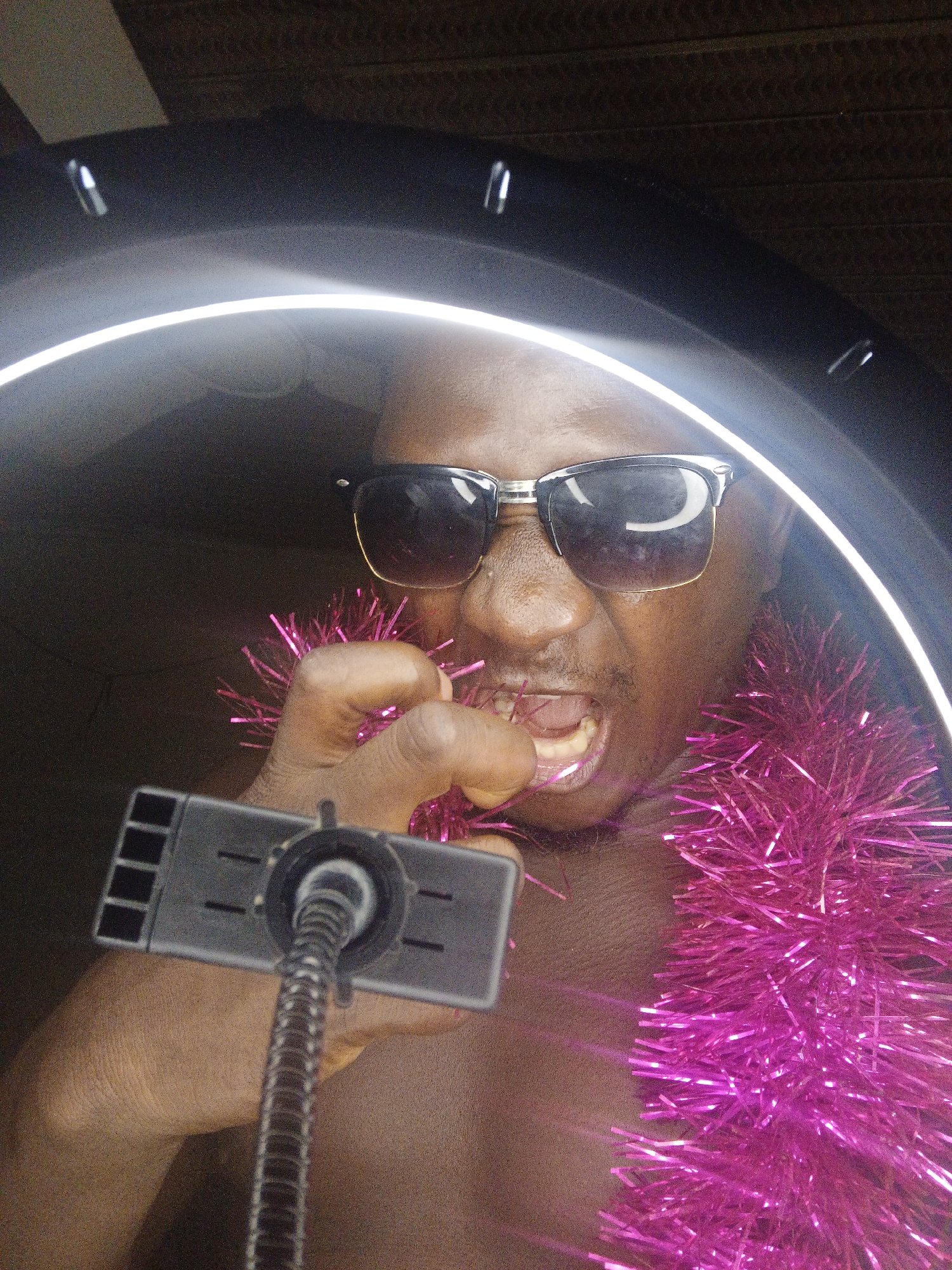A medium shot captures a person from the chest up, inside a ring light. The person, wearing sunglasses and bright pink tinsel around their neck, has their mouth open and is holding a metallic object up to their face with their left hand. The ring light is on and is a prominent light source in the image. The background appears to be dark and textured, possibly a wooden ceiling. The overall scene suggests a close-up, candid moment captured with artificial lighting.