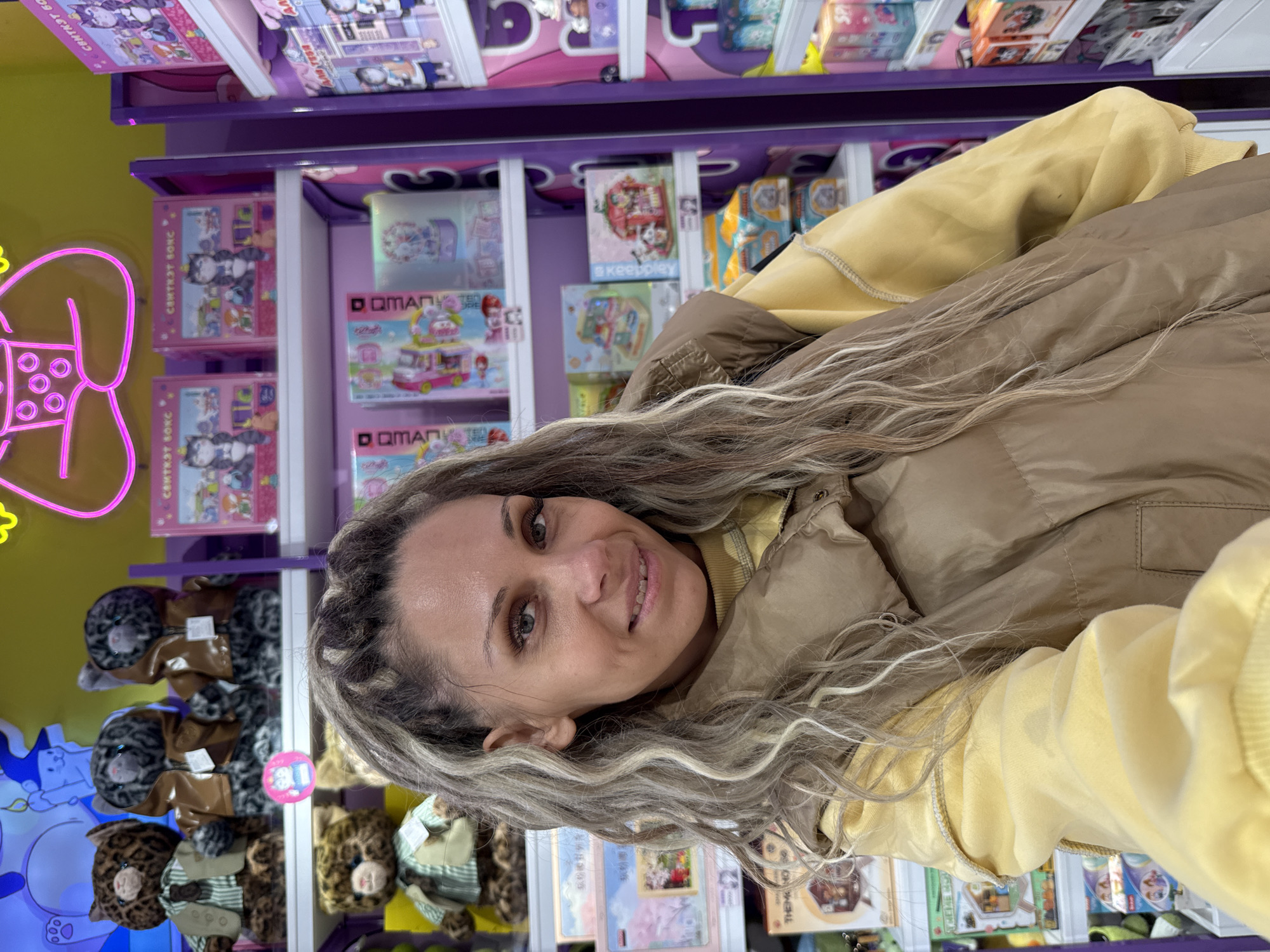 In Yaroslavl, Russia, a woman with long, blonde-streaked dreadlocks is taking a selfie in what appears to be a toy store. She is wearing a yellow hoodie and a tan puffer vest. Behind her, shelves are stocked with various toys, including stuffed animals and boxed play sets. The shelves are illuminated by bright overhead lighting and a neon sign that spells out a word in Russian. The woman is smiling at the camera.
