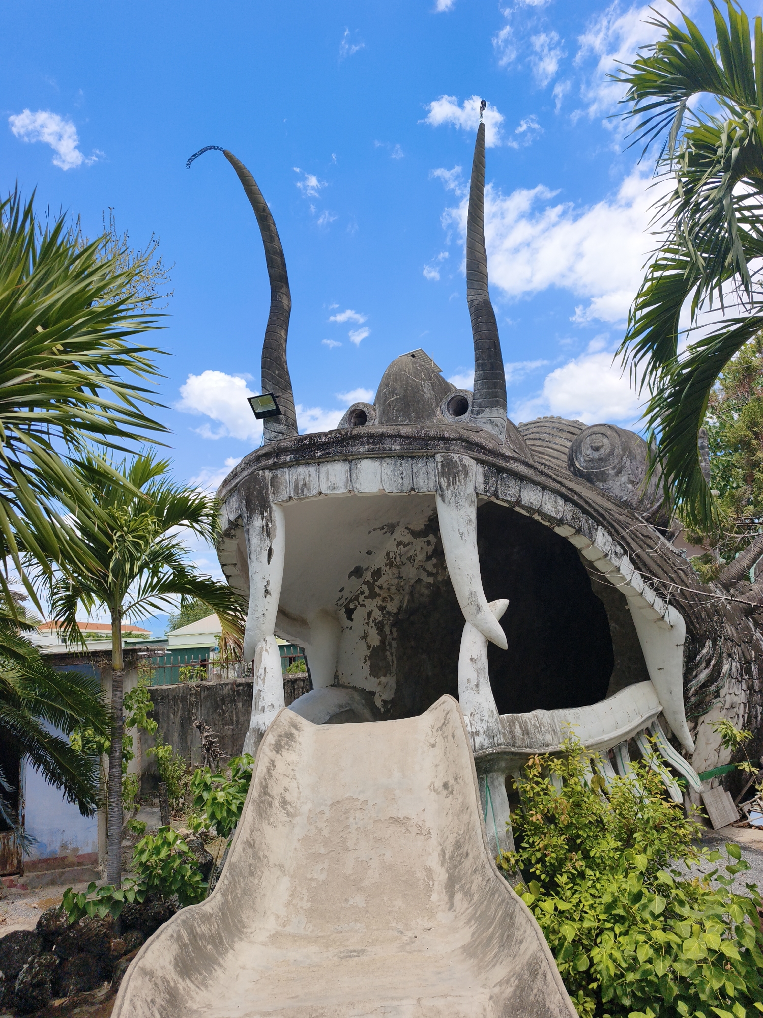 The image depicts a concrete sculpture of a monster's head, likely a dragon or a similar mythical creature, with its mouth open to reveal a slide. The sculpture is weathered and appears to be part of an outdoor attraction or park. Palm trees and other lush greenery surround the structure, and a bright blue sky with scattered white clouds forms the backdrop. In the distance, buildings and a fence are visible, suggesting an urban or semi-urban environment. This landmark is located in Cam Ranh, Vietnam, specifically within the Buddha Park, also known as Tinh Xa Ngoc Son. The park is famous for its numerous Buddhist statues and sculptures, including this impressive, albeit somewhat decaying, monster head slide.