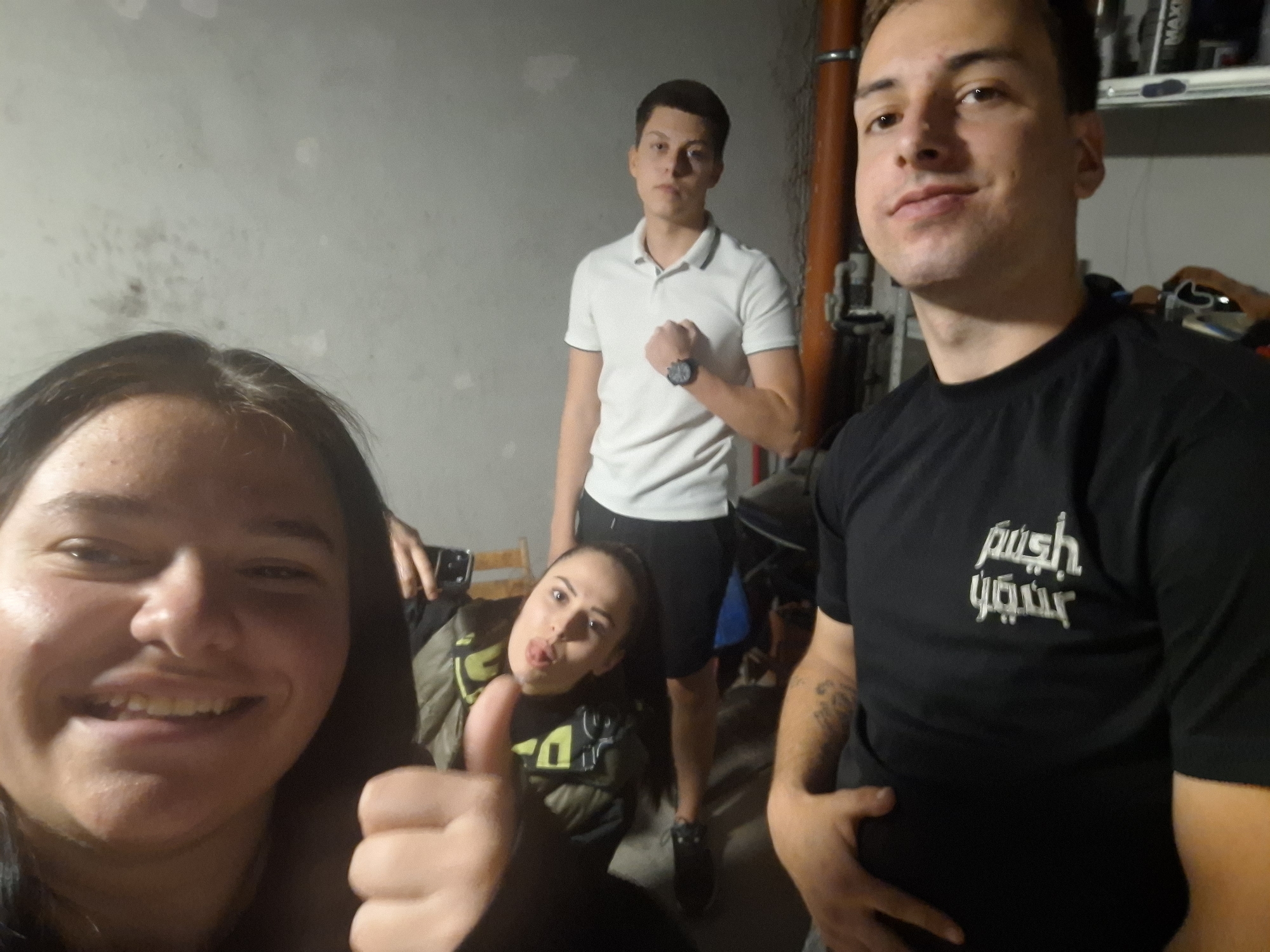 In an indoor setting, potentially a basement, five individuals are captured in a seemingly impromptu photograph. One person, presumably holding the camera, is in the foreground on the left, smiling broadly and giving a thumbs-up. Behind this person, another individual is seated, making a face with their tongue slightly protruding. In the background, a person stands wearing a white polo shirt and dark shorts. To the right and slightly forward of the first person, another individual is visible, wearing a black t-shirt. The final person is partially visible in the background. The environment includes a plain, possibly concrete wall and assorted items.