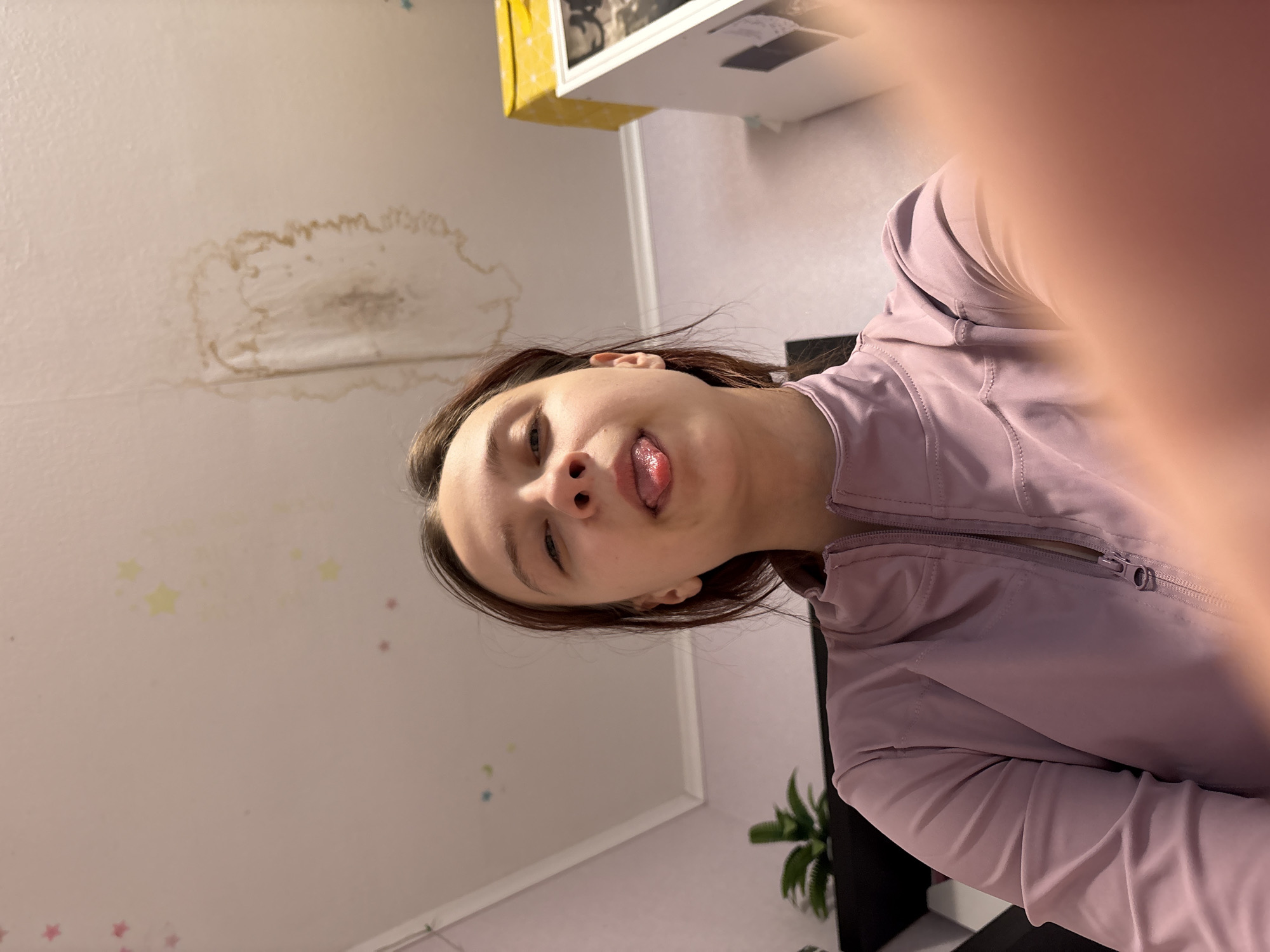 A tilted, close-up selfie features a young woman with fair skin and dark hair sticking out her tongue, her left eye partially closed. She is wearing a pink athletic jacket with a visible zipper. The background includes a white wall with a discolored water stain near the top left corner, and small star-shaped decals scattered on the lower half of the wall. A white shelf is visible in the upper right, along with a small houseplant located in the bottom right corner. The location is presumably Прикамский, Russia.