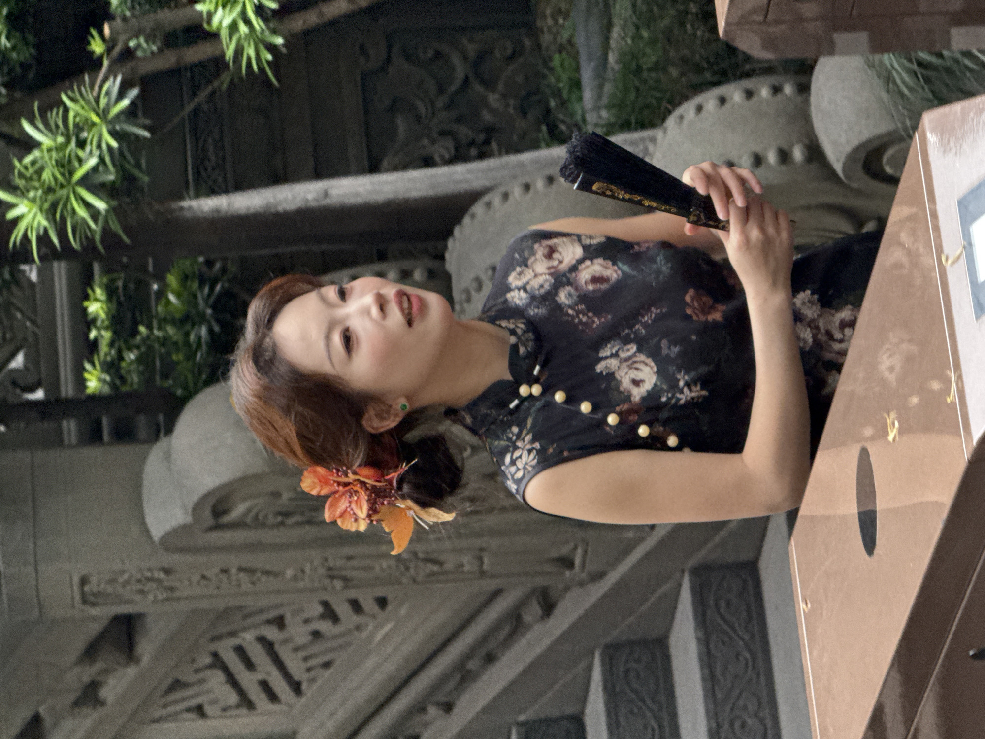 A woman is holding a black fan in an outdoor setting. The location appears to be a park-like area, possibly a traditional garden, within the Yuzhong District, China. The woman wears a dark floral patterned dress with buttons on the side. Her hair is styled with an orange flower accessory. Architectural elements with intricate carvings are visible in the background.