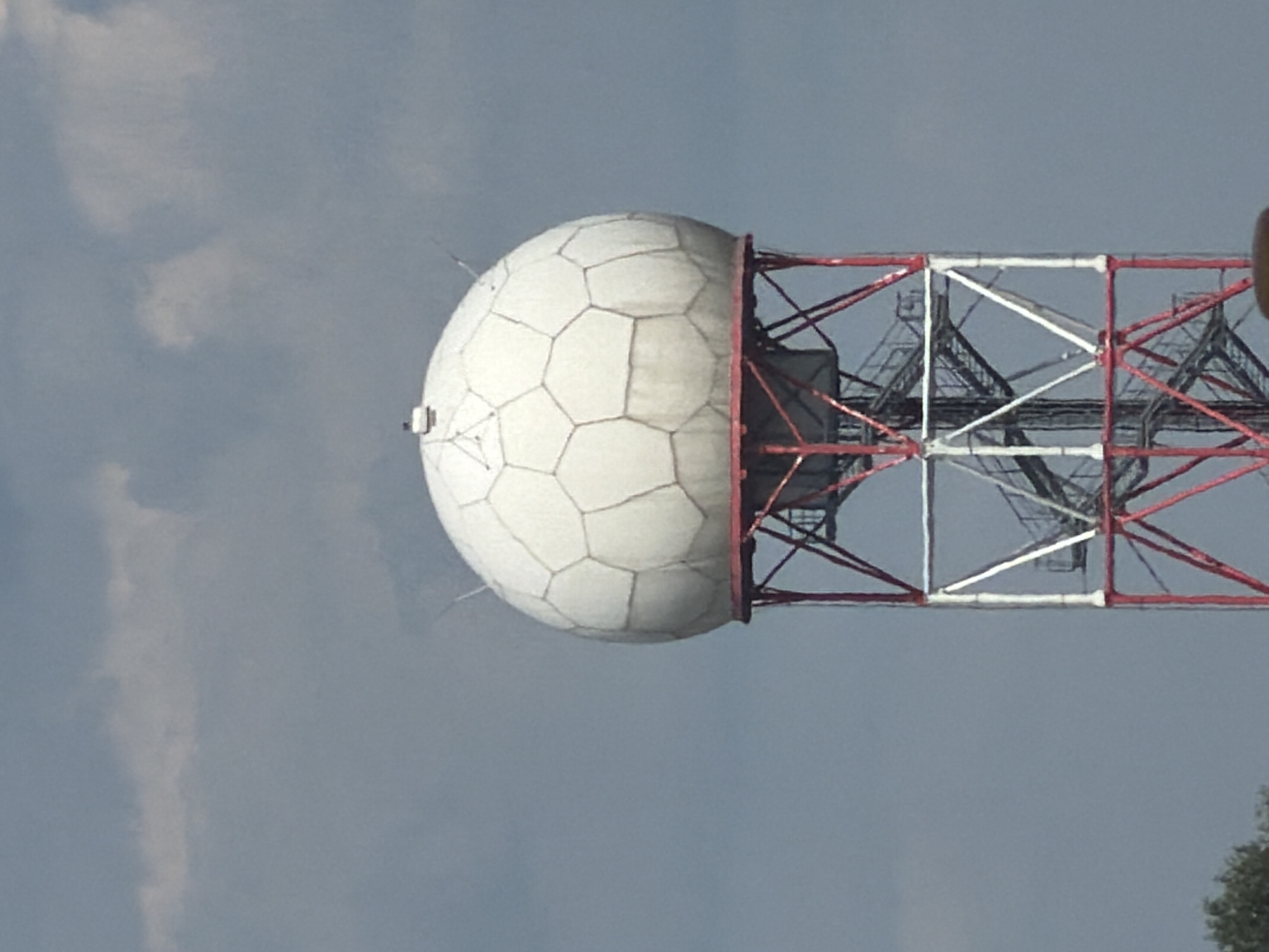 The image shows a close-up, eye-level, outdoor shot of a weather radar dome mounted on a tall, red and white lattice tower. The dome is white and appears to be made of hexagonal panels. The sky behind the dome is overcast with gray clouds. A portion of trees is visible on the bottom left edge of the frame. The scene appears to be located in an open area, possibly on the outskirts of a city. This particular weather radar is located at the National Institute of Meteorology and Hydrology of the Bulgarian Academy of Sciences in Sofia, Bulgaria.