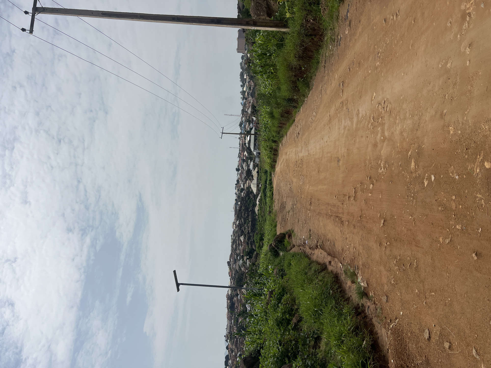 The image depicts a dirt road leading through a densely populated area in Jos, Nigeria. The road is reddish-brown and appears to be unpaved, with visible stones and tire tracks. On either side of the road, there is lush green vegetation, including what appears to be corn stalks and other plants. In the background, a sprawling settlement covers a hilly landscape, with numerous buildings constructed from various materials, mostly with corrugated metal roofs. Power lines stretch across the sky, supported by wooden poles. A street light is visible on the left side of the road, and a larger, more modern building can be seen in the distance on the right, suggesting a mix of development within the settlement. The sky is overcast with scattered clouds.