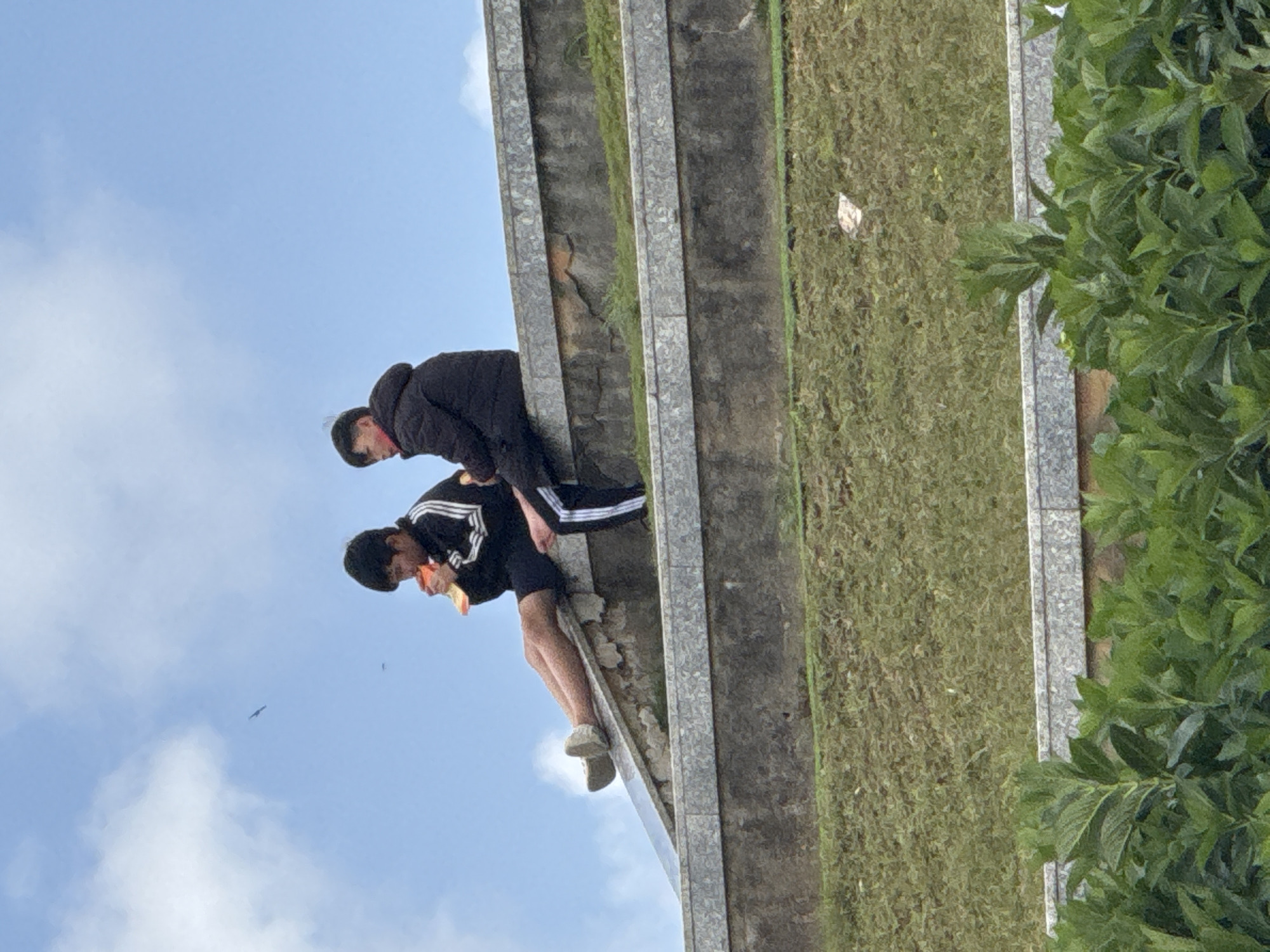 In Da Lat, Vietnam, two young individuals are seated on a stepped concrete structure, with grassy slopes below. The person on the left, facing right, is wearing a black jacket with white stripes and dark shorts. They are holding a piece of orange food and appear to be eating. Their legs are extended forward, and they are wearing light-colored shoes. To their right, the second individual is wearing a dark puffer jacket and dark pants with white stripes down the legs. They are facing left and looking downwards. The sky above is blue with scattered white clouds, and a bird is visible in flight in the distance. The concrete structure has a stone-like finish on its edges, and some vegetation grows on the grassy slopes.