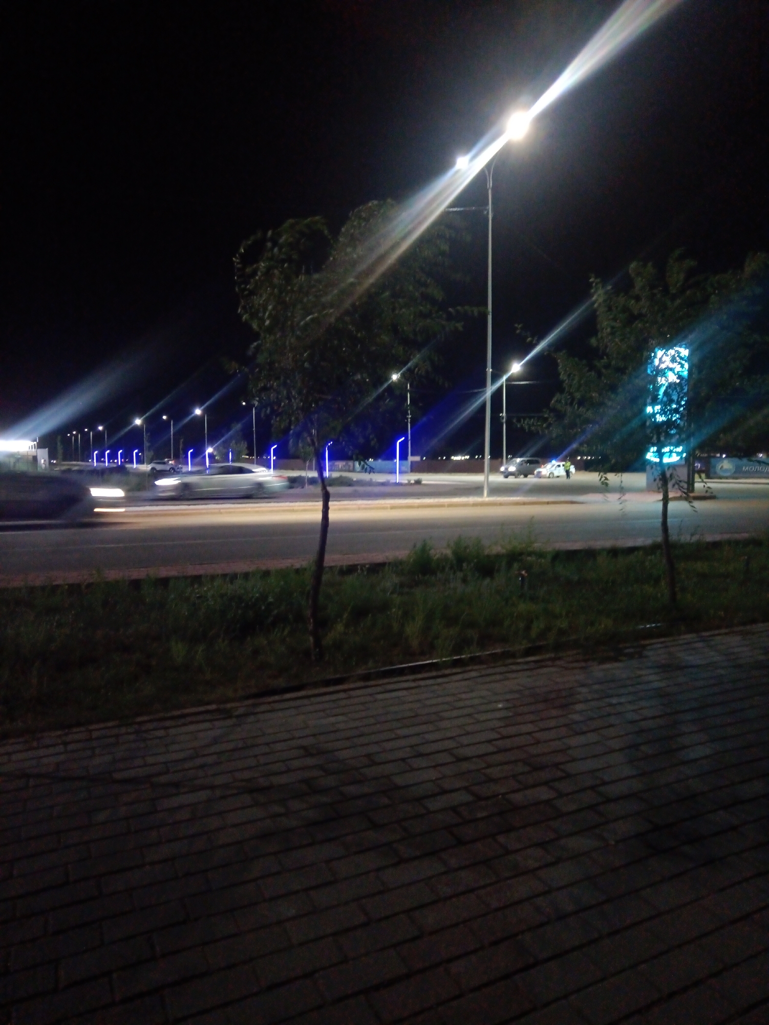This is a nighttime photograph taken from a sidewalk, looking towards a road and a brightly lit area beyond. The foreground is dominated by a textured brick walkway, casting long shadows. Beyond the walkway is a strip of dark grass and then a paved road. Several cars are visible on the road, some in motion, indicated by motion blur. A streetlamp with a bright light illuminates the scene, creating lens flares and light streaks across the image. Trees line the side of the road. In the background, there are more streetlights, some emitting a bluish light. There is also a large, brightly lit sign with a blue and white pattern, possibly an advertisement or information display. The scene appears to be an urban or suburban setting in Balqash, Kazakhstan, likely a public space or commercial area illuminated for nighttime activity.