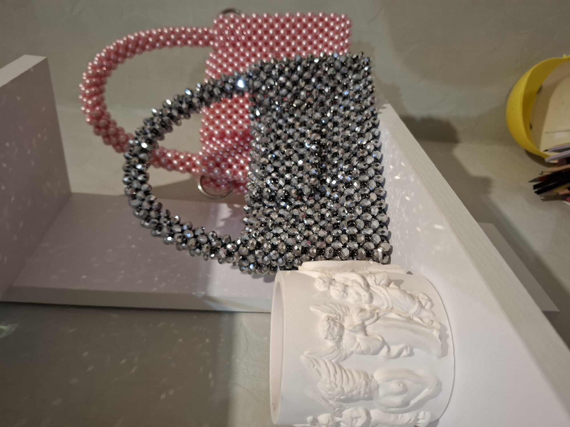 The image displays two beaded handbags and a decorative ceramic pot with angel figures, positioned on a white shelf. The handbags are one pink and one silver, both featuring intricate beadwork and handle designs. The ceramic pot is white and adorned with relief carvings of angels. The scene appears to be an indoor setting, possibly a display shelf in a home or store. There is no indication of the city of Korenovsk, Russia.