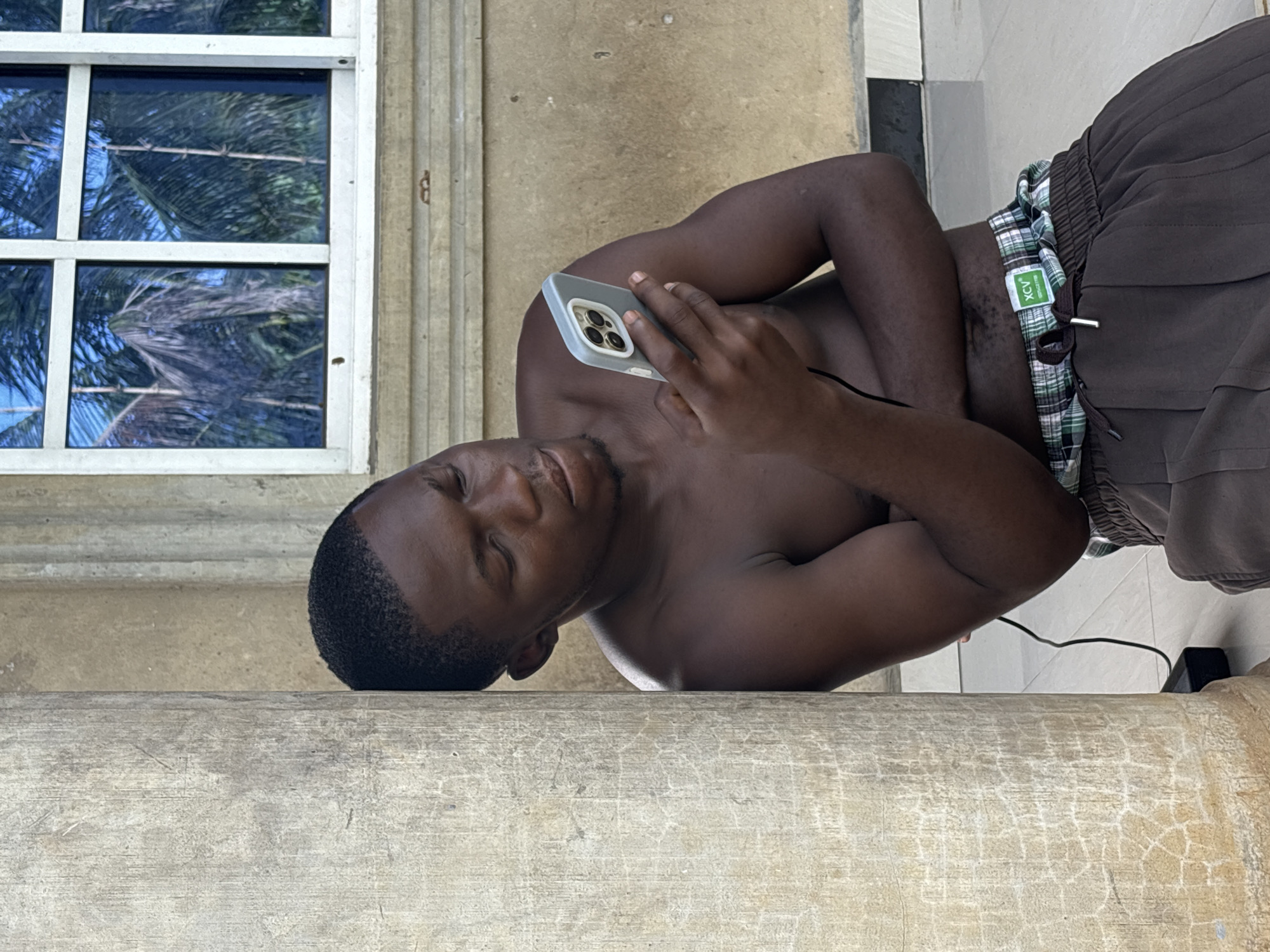 A shirtless Black male is lying on a ledge and holding a smartphone in his right hand, in Ikot Ekpene, Nigeria. He is wearing dark-colored shorts with a green and white patterned waistband. A window with blue glass panes is visible to the left, reflecting palm trees. The ledge is light gray with a weathered surface. A black charging cord is visible, connected to the phone.