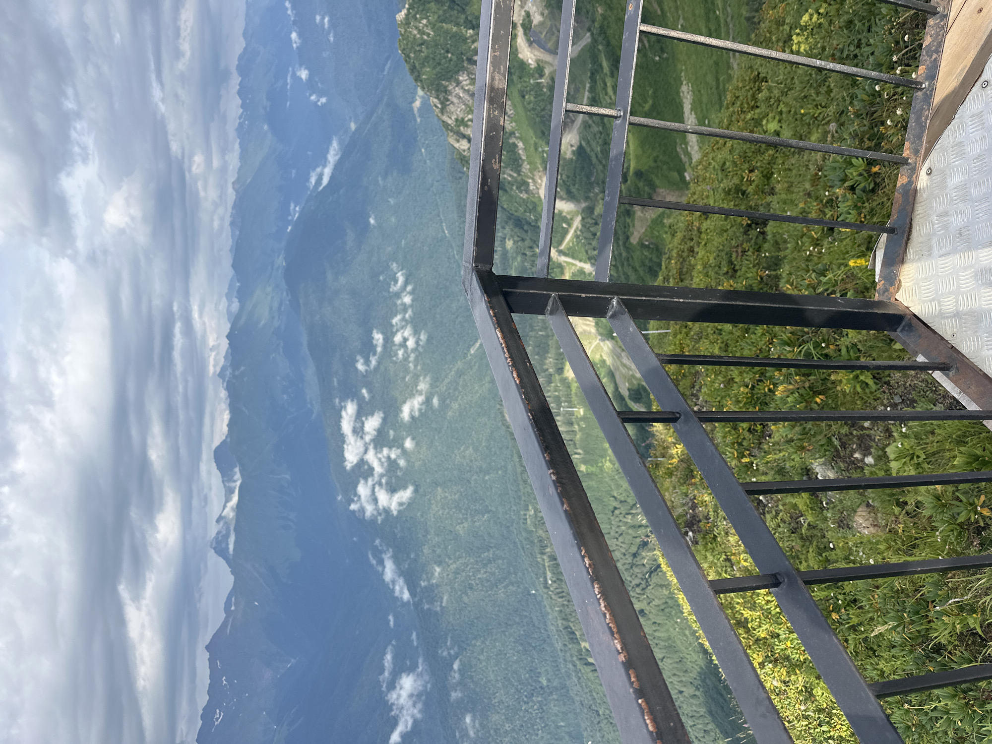The image captures a scenic mountain vista from an elevated viewpoint in Estosadok, Russia. In the foreground, a black metal railing with visible signs of rust borders a metal platform. Beyond the railing, lush green slopes dotted with yellow flowers descend into a valley. In the middle ground, a winding road can be seen cutting through the greenery. The background is dominated by a series of majestic, layered mountain ranges under a cloudy sky. Patches of white clouds drift between the peaks, adding depth and texture to the landscape. The overall impression is one of natural beauty and elevation, suggesting a location offering panoramic views.