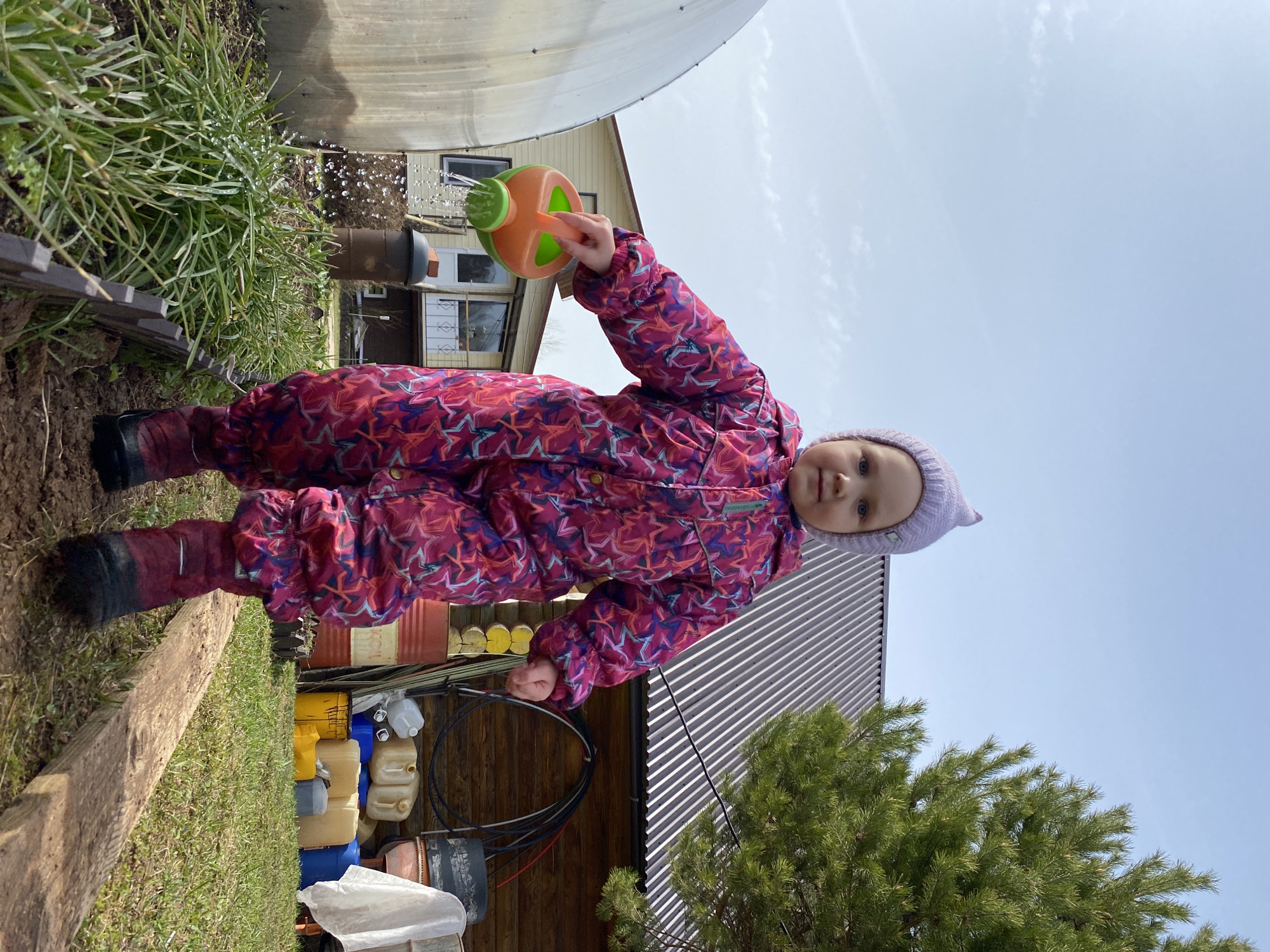 A young child, likely a toddler, is captured in a full-length outdoor portrait, engaged in watering plants or the ground. The child wears a vibrant, patterned full-body suit with stars in shades of pink, purple, and blue, and a light purple knitted hat. They hold an orange and green plastic watering can with both hands, from which water is actively pouring. The child looks directly at the camera with a neutral to slightly curious expression.
The setting appears to be a backyard or garden in Фоки, Russia, under a bright, clear daytime sky. Lush green grass and other foliage are prominent in the foreground and mid-ground, with visible water droplets on some leaves. A rough wooden beam or plank runs along the lower left foreground.
In the background, a large, translucent greenhouse or similar covered structure is visible on the left. Behind it, a house or shed with windows can be partially seen. To the right, various colorful containers, drums, and other supplies are stacked near a wooden structure. A green conifer tree stands on the far right of the frame. The scene suggests a pleasant outdoor activity on a clear day. No visible text is present in the image.