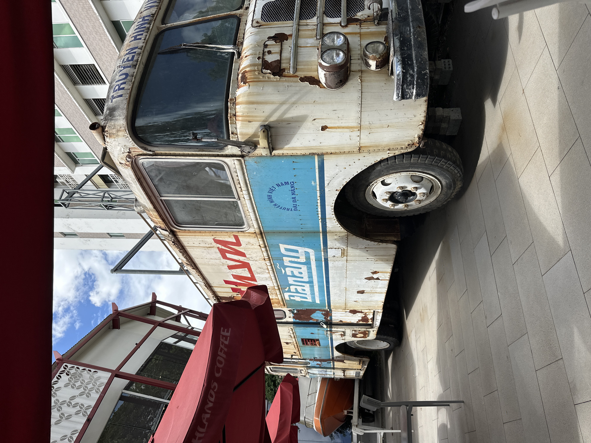 The image displays a weathered, vintage bus, likely repurposed as a cafe or food truck, parked on a paved area in Đà Nẵng, Vietnam. The bus is predominantly white with a prominent blue stripe and some red lettering, showing considerable rust, peeling paint, and general wear. Its front features large, circular headlights and a distressed grille.
Along the blue stripe on the side of the bus, the text "TRUYỀN HÌNH VIỆT NAM" (Vietnamese Television) and "ĐÀ NẴNG" are clearly visible. The phrase "TRUYỀN HÌNH VIỆT NAM" is also partially discernible across the front top of the bus. Red lettering, with "Mỹ" being clear, is painted above the blue stripe. In the foreground, several open red parasols are visible, one prominently displaying "HIGHLANDS COFFEE," suggesting an outdoor seating area associated with the bus or a nearby establishment.
The bus rests on a light-colored, rectangular tiled pavement. In the background, a modern, multi-story building with a pattern of light and dark panels and green-tinted windows rises. To the lower left, partially obscured by the parasols, is another building characterized by a white facade and a distinct red-framed roof structure. Patches of blue sky with white clouds are visible between the buildings.
The scene takes place during daytime under sunny conditions, as indicated by the clear lighting and visible shadows. There are no people present, and the setting appears to be static. The vintage bus, with its historical markings and current state of charming disrepair, stands out against the more modern architectural backdrop, creating a unique visual blend in an urban Vietnamese context.