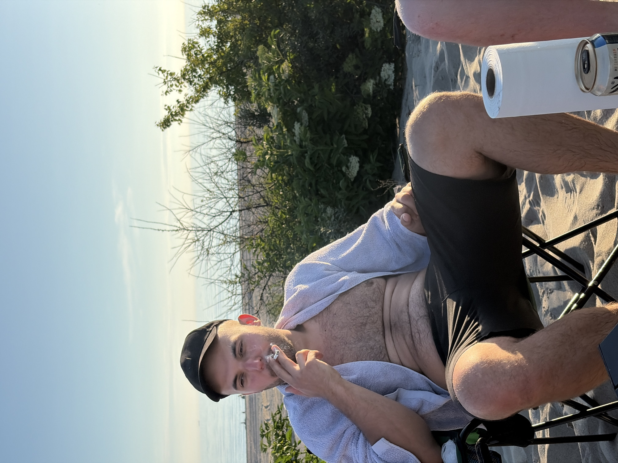 A man wearing a black baseball cap and a white towel draped over his shoulders sits in a folding chair on a sandy beach. He is smoking a cigarette and looking to his left. He is wearing dark shorts. In front of him is a roll of paper towels and a can. Behind him are some bushes and a body of water. The sky is clear and blue, with a faint hint of clouds. The location is identified as Povarovka, Russia.
