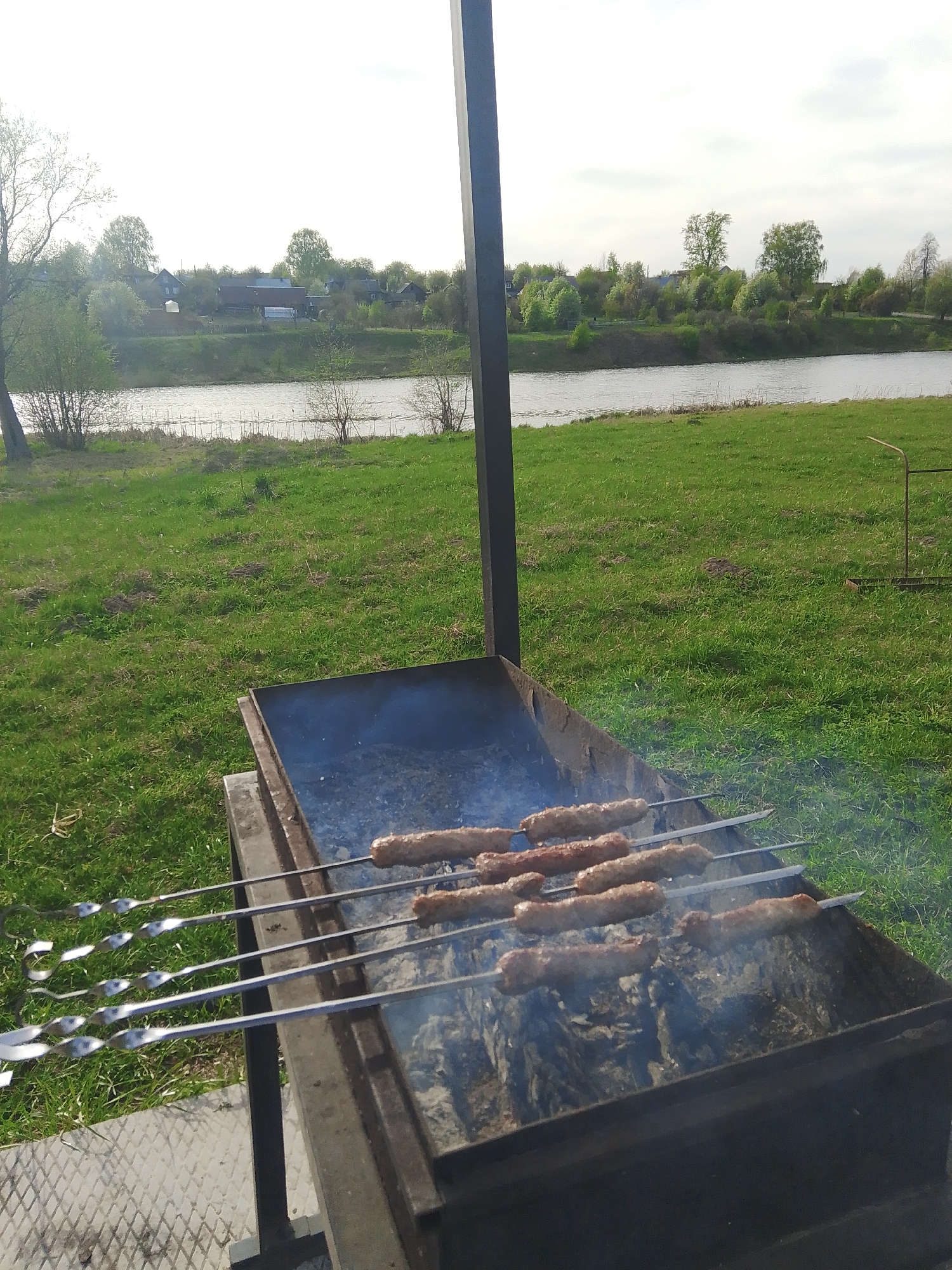 A medium, eye-level shot shows several metal skewers laden with sausage-like meat roasting on a barbecue grill set up in a grassy area, potentially in Фоки, Russia. In the background, a body of water can be seen, with houses and trees on the opposite bank. The grill is black and rectangular, with visible smoke rising from the charcoal. The skewers extend beyond the grill on both sides. The ground is covered in green grass, and there is a partially visible metal structure above the grill. The sky is overcast, suggesting a cool day.