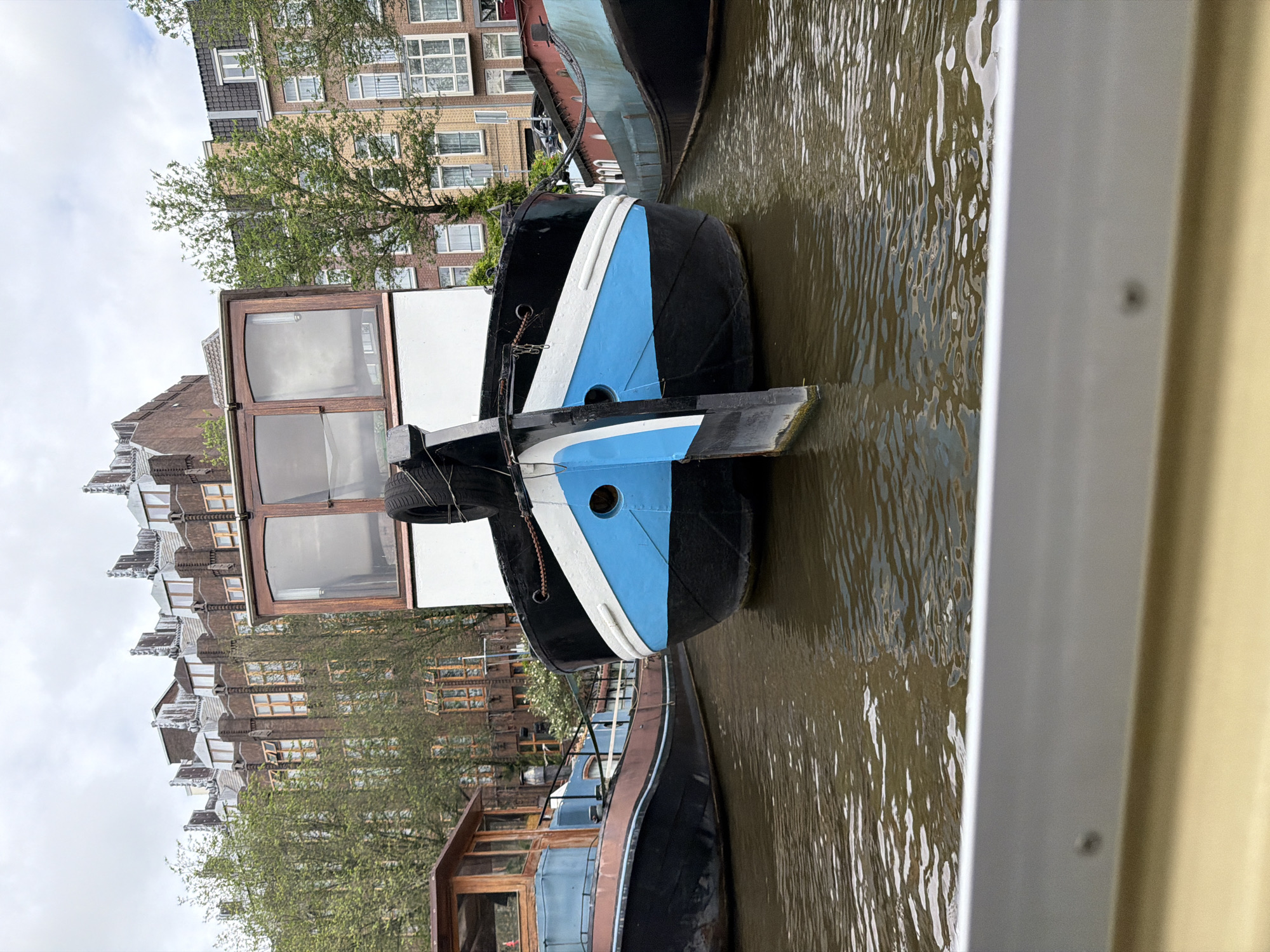 A low-angle, medium shot captures the front of a black and blue canal boat docked in Amsterdam, Netherlands. The boat has a white cabin with wooden window frames and a spare tire mounted on the bow. Two other canal boats are visible to the left and right of the main subject. In the background, multi-story buildings with distinctive Dutch architecture line the canal. Trees with green foliage are interspersed between the buildings. The sky is overcast. The water in the canal is a murky brown. A sliver of a light-colored railing or boat in the foreground partially obscures the bottom of the frame.