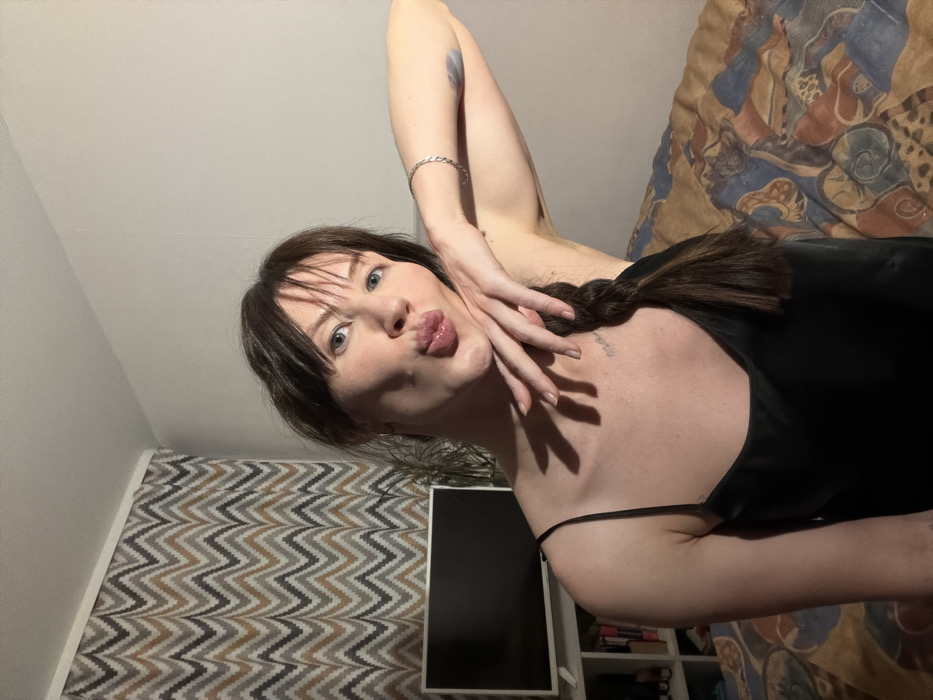 The scene is located in a residential setting within Perm, Russia, depicting a female individual from the mid-chest upwards. She possesses dark brown hair styled with bangs and a visible braid extending over her left shoulder. Her face is directed towards the viewer with blue eyes, and her lips are pursed in a "kiss" gesture. Her left hand is positioned with fingers splayed against her chin and lower jawline, casting shadows onto her upper chest. A small, dark-ink text tattoo, resembling "Matthew," is visible on her upper right chest near the clavicle. Her right arm is bent at the elbow, revealing a silver-colored braided bracelet on her wrist and a light blue abstract tattoo on her upper right bicep. She is wearing a black, thin-strapped top, likely made of satin or silk material.
In the background to the left, a dark-screen flat-panel television rests on a white shelf unit. Below the television, a white shelving unit holds various objects, including books. A green-leafed potted plant is situated between the woman and the television. The wall immediately behind the subject is plain and light-colored, transitioning to a patterned wall covering featuring a brown, grey, and cream chevron design on the far left. The ceiling is plain white. The woman is seated or reclined on a surface, likely a bed or sofa, covered by a patterned textile that displays abstract shapes in shades of brown, orange, blue, and cream.