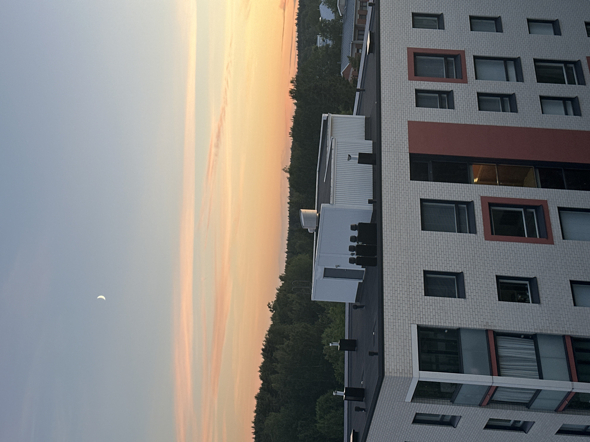 An elevated view captures a serene twilight scene above a city. The sky transitions from a soft blue at the top to a warm orange and pink gradient near the horizon, adorned with delicate streaks of clouds. A thin crescent moon is visible in the upper left quadrant of the sky. Below the sky, a dense, dark green forest forms a horizontal line across the mid-ground.
In the foreground, the rooftops of modern buildings are prominent. The most detailed building in the foreground is a multi-story residential complex with light-colored brickwork. Its facade features numerous windows of varying sizes, some with dark frames and others with distinctive reddish-orange accents. Several balconies with glass railings and sheer curtains are visible.
On the rooftops, various structures are present, including ventilation systems, pipes, and a white, boxy building with a door and corrugated metal siding. The overall atmosphere is tranquil and urban, with a natural element provided by the distant forest and the celestial bodies.