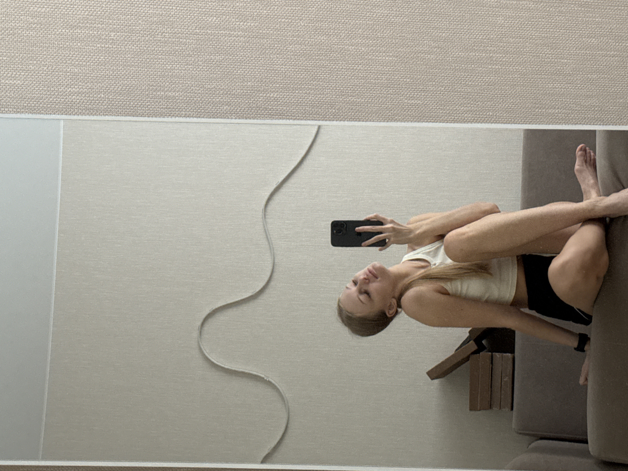 A young woman with blonde hair, wearing a white crop top and black shorts, is taking a mirror selfie in what appears to be an apartment. She is sitting on a sofa with her legs crossed and her head tilted back, holding a smartphone to capture the reflection. In the background, there is a textured wall with a wavy decorative element and a stack of brown boxes on a piece of furniture. The overall environment suggests a casual, indoor setting. The image does not provide any specific indicators to suggest the location is in the city of Perm, Russia.