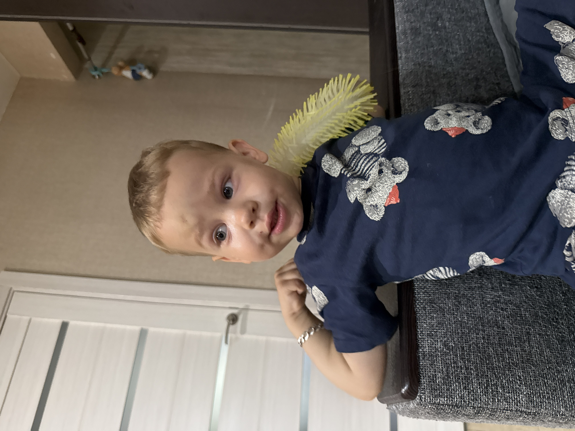 A young boy with blond hair and blue eyes is seated on a couch. He is wearing a dark blue t-shirt with a pattern of grey teddy bears. A yellow, spiky toy is positioned behind his neck. The boy has a bracelet on his left wrist. To the left of the boy is a white door with horizontal panels and a silver handle. Behind the boy, the wall is a neutral beige color, with a dark brown wardrobe to the right. A small doll or toy is hanging from a string on the wall in the background. The setting appears to be indoors, likely a room in a house or apartment. The image is taken from a slightly low angle.