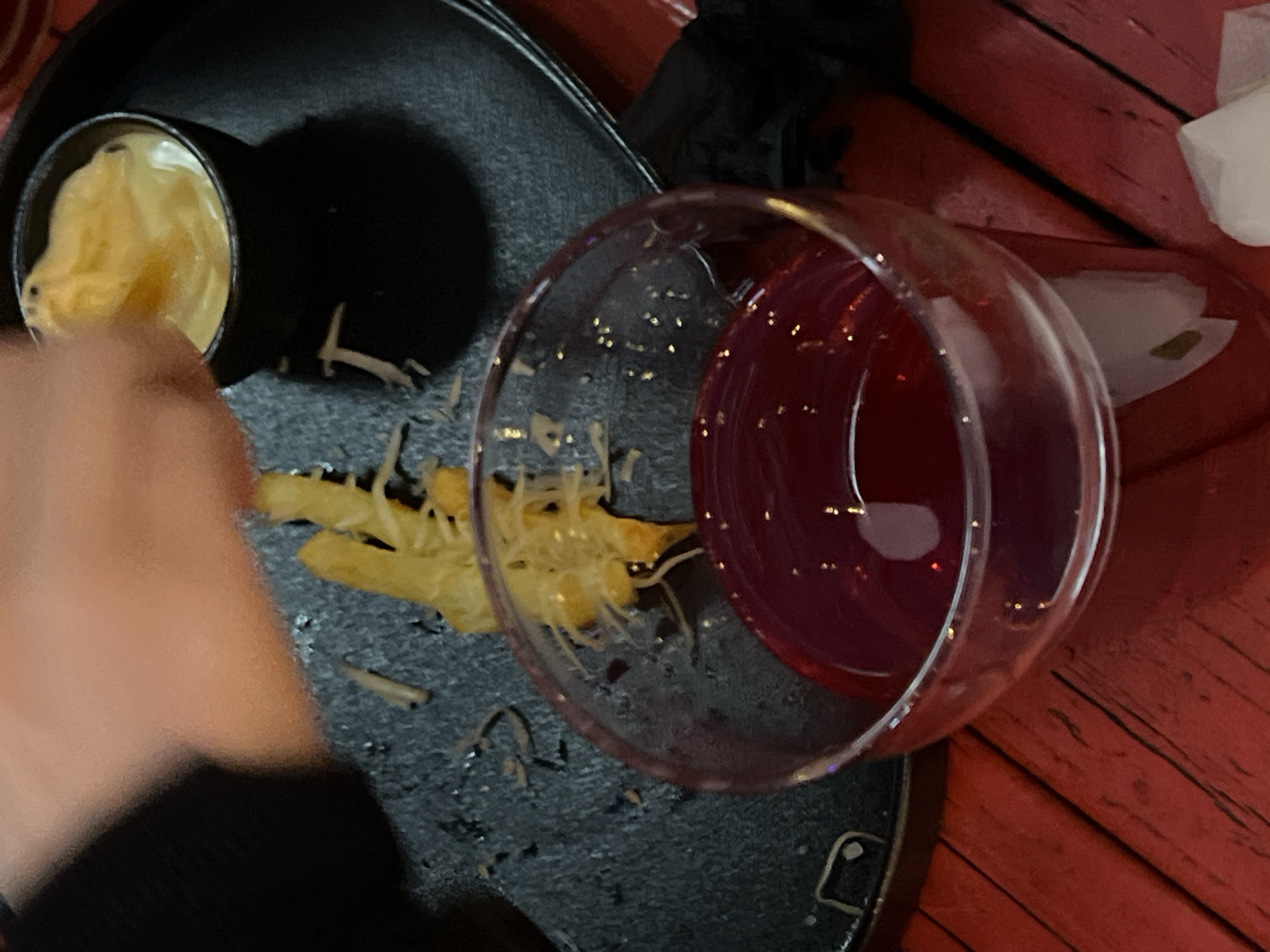 The scene shows a black tray holding a small, black ramekin filled with a yellow sauce, alongside scattered french fries. A wine glass containing a red liquid is present on the tray. A portion of a person's arm and black clothing is visible on the left side of the frame, suggesting someone is reaching for the food. The background reveals a red, wooden surface. The location and mention of Vologda, Russia is inconsistent with the visual elements of the provided scene. The scene seems to be depicting a meal at a restaurant or similar establishment.