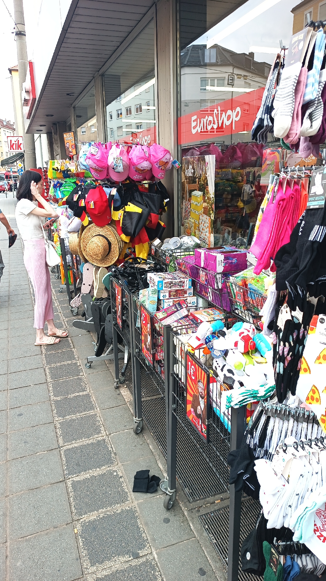 The image depicts a street-level view of a Euroshop store in Nuremberg, Germany. A woman, wearing a white t-shirt and pink pants, stands by a display of hats and bags, looking at her phone. In front of the store, racks of merchandise are set up on the sidewalk, including pink children's hats, black and yellow German flag-themed bags, straw hats, and various colorful socks and other small items. The Euroshop store has large glass windows displaying more merchandise inside. The sidewalk is paved with gray square tiles. Two pairs of black socks are on the ground in front of the merchandise racks. The overall scene suggests a bustling commercial area.