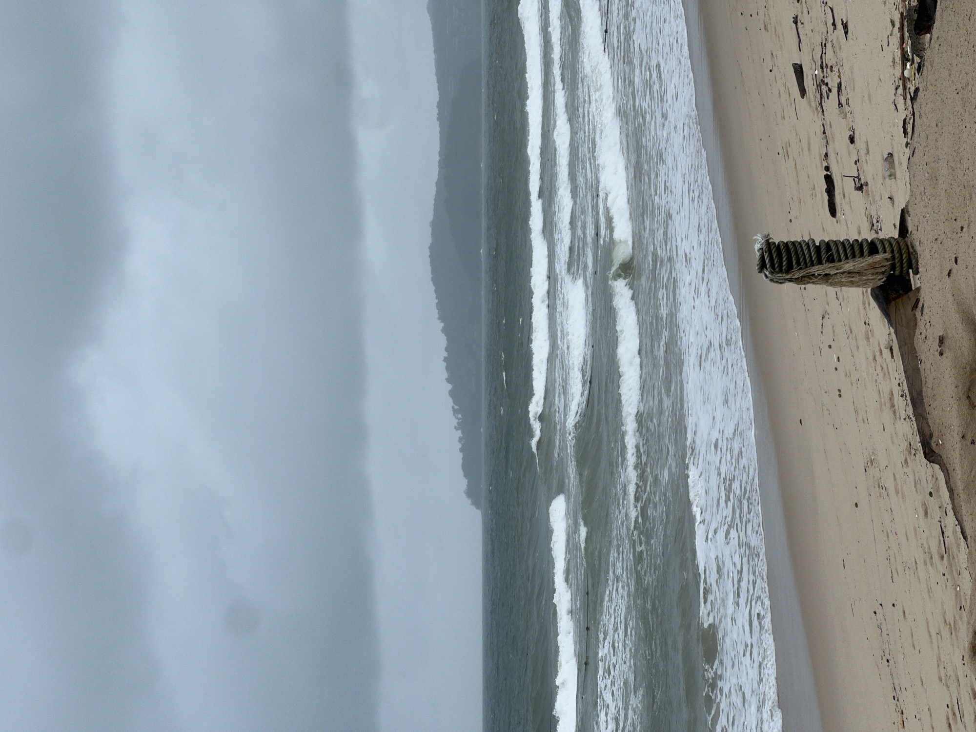 The image displays a coastal scene on an overcast day, oriented horizontally with the sky at the top, ocean in the middle, and beach on the right. The sky is entirely covered with thick, light grey clouds, creating a muted and hazy atmosphere.
The central part of the image features the ocean, with several lines of white-capped waves breaking and rolling towards the shore. The water appears a greenish-grey hue, reflecting the somber sky. In the distance, a low-lying coastline, possibly hills or mountains covered in vegetation, is visible through the mist or clouds, appearing as a dark, indistinct silhouette.
To the right, a sandy beach stretches along the coastline. The sand is light brown and appears somewhat damp near the waterline. A notable man-made object is positioned on the beach in the foreground: a sturdy wooden post or groyne, heavily wrapped with thick, braided rope. The top of the rope appears frayed. Small dark debris is scattered on the sand around this structure, suggesting natural detritus or litter. No specific human activity is visible, and there is no visible text. The overall impression is one of a calm, perhaps slightly melancholic, day at the beach under a heavily clouded sky.