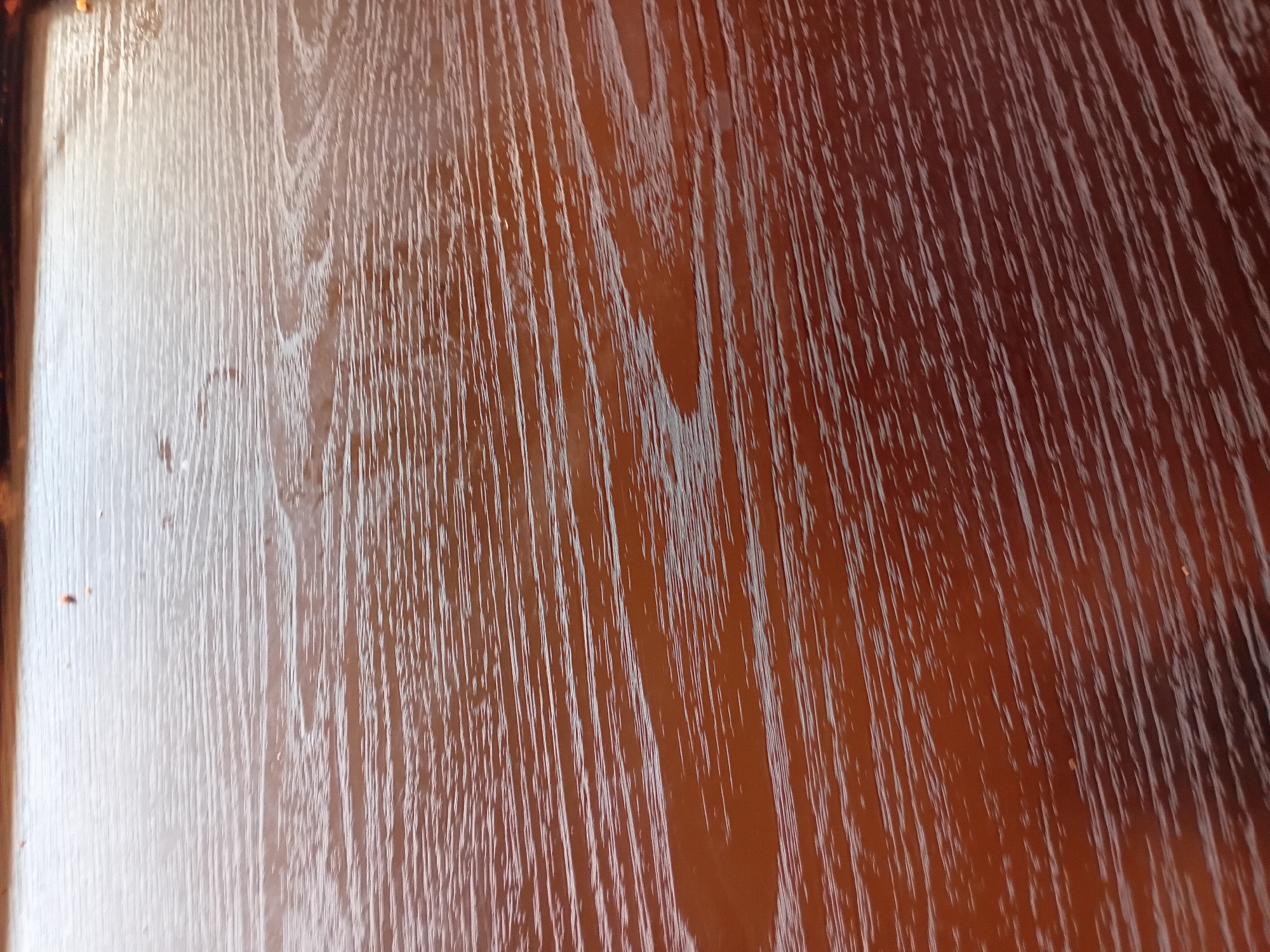 The image shows a close-up of a polished wooden surface. The wood grain is visible as parallel, wavy lines of lighter brown against a darker brown background. The surface is highly reflective, with bright streaks of light that highlight the texture and sheen of the wood. There are a few small, dark specks on the surface, possibly dust or debris. The perspective is from above, looking down at an angle. The scene appears to be indoors, given the controlled lighting. The provided location information for Howayi, Nigeria, is not directly discernible from the image itself, as there are no environmental cues such as buildings, landmarks, or specific flora that would indicate this location.