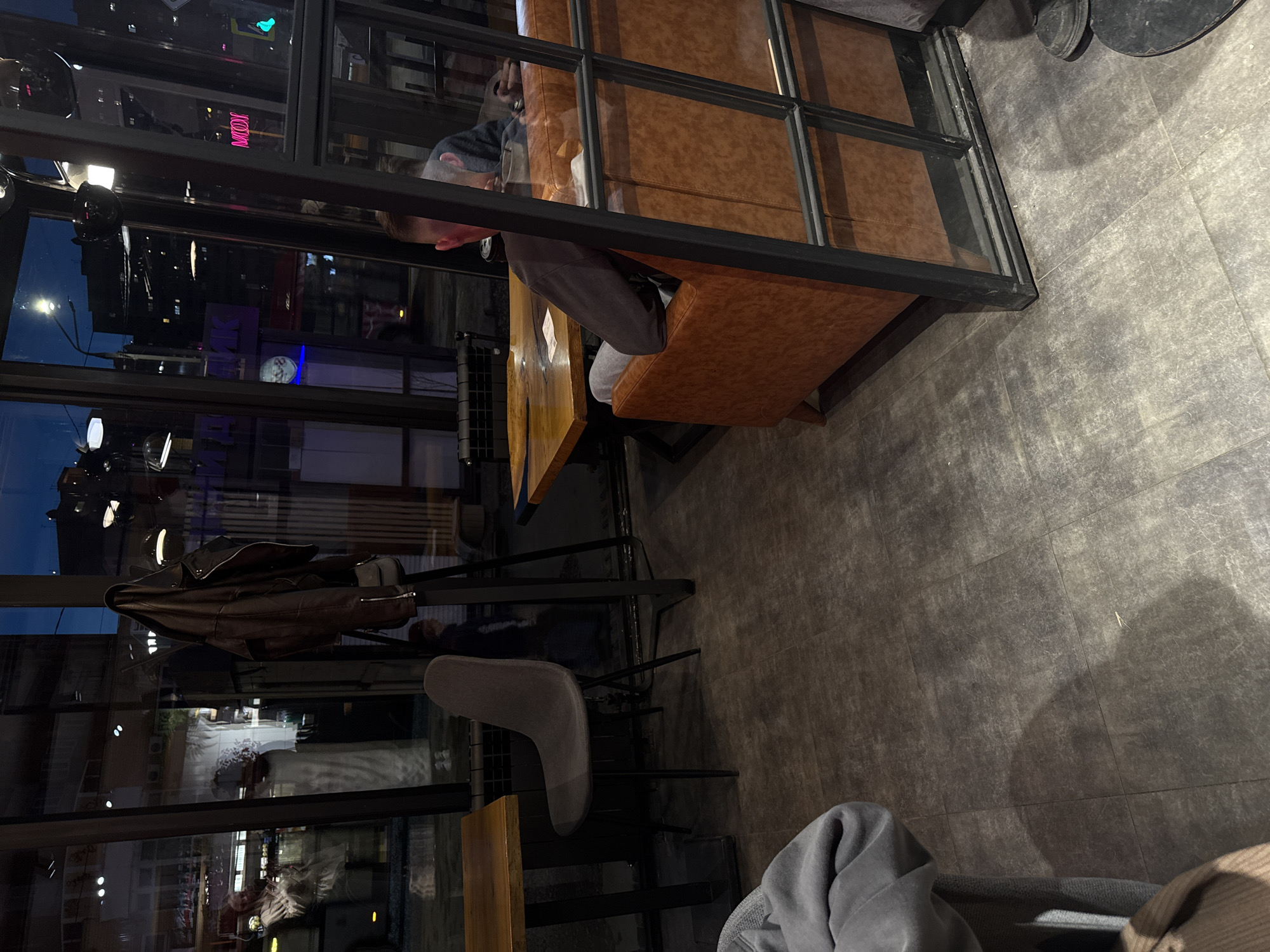 The image depicts an interior scene, likely a restaurant or cafe, located in Perm, Russia, judging by the reflections of buildings and streetlights visible through the large glass windows.
The foreground features a seating area with brown upholstered booth seating enclosed by a black metal frame with glass panes. A man is seated within this booth. The reflections of a city street with buildings and streetlights are visible in the glass of the seating area.
A leather jacket is hanging near the seating area. A gray chair is partially visible near a wooden table. The floor is covered in gray tiles.
Additional patrons or staff members are not immediately apparent within the view, and the overall environment suggests a modern or contemporary design aesthetic.