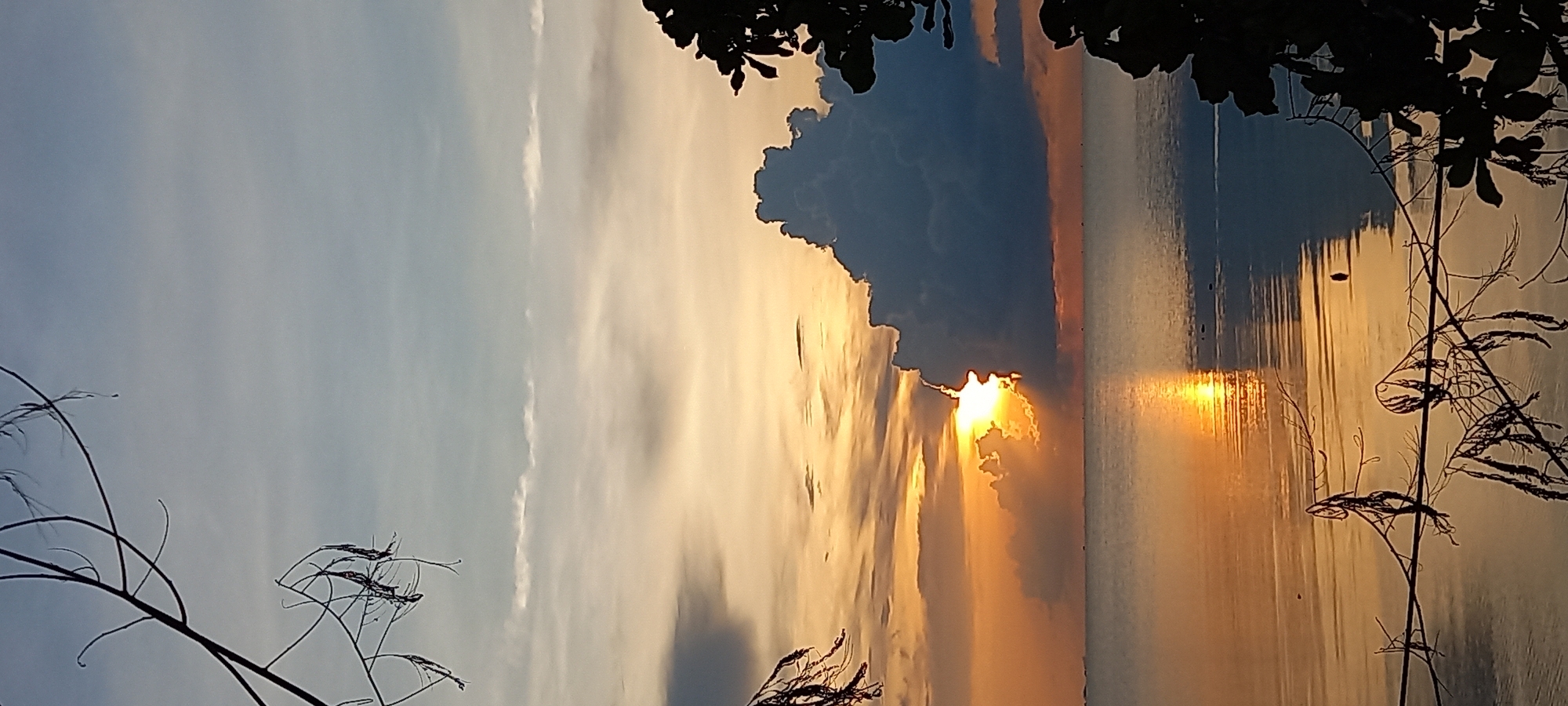 The image depicts a sunset over water, likely the sea, as seen from Baan Wok Tum, Thailand. The sun is partially obscured by a large, dark cloud, creating a dramatic effect with visible sun rays. The water reflects the golden and orange hues of the sunset. Silhouetted tree branches frame the scene at the top and along the right edge, with some thin branches also visible on the lower left. The sky above the clouds has a gradient from pale blue to orange.