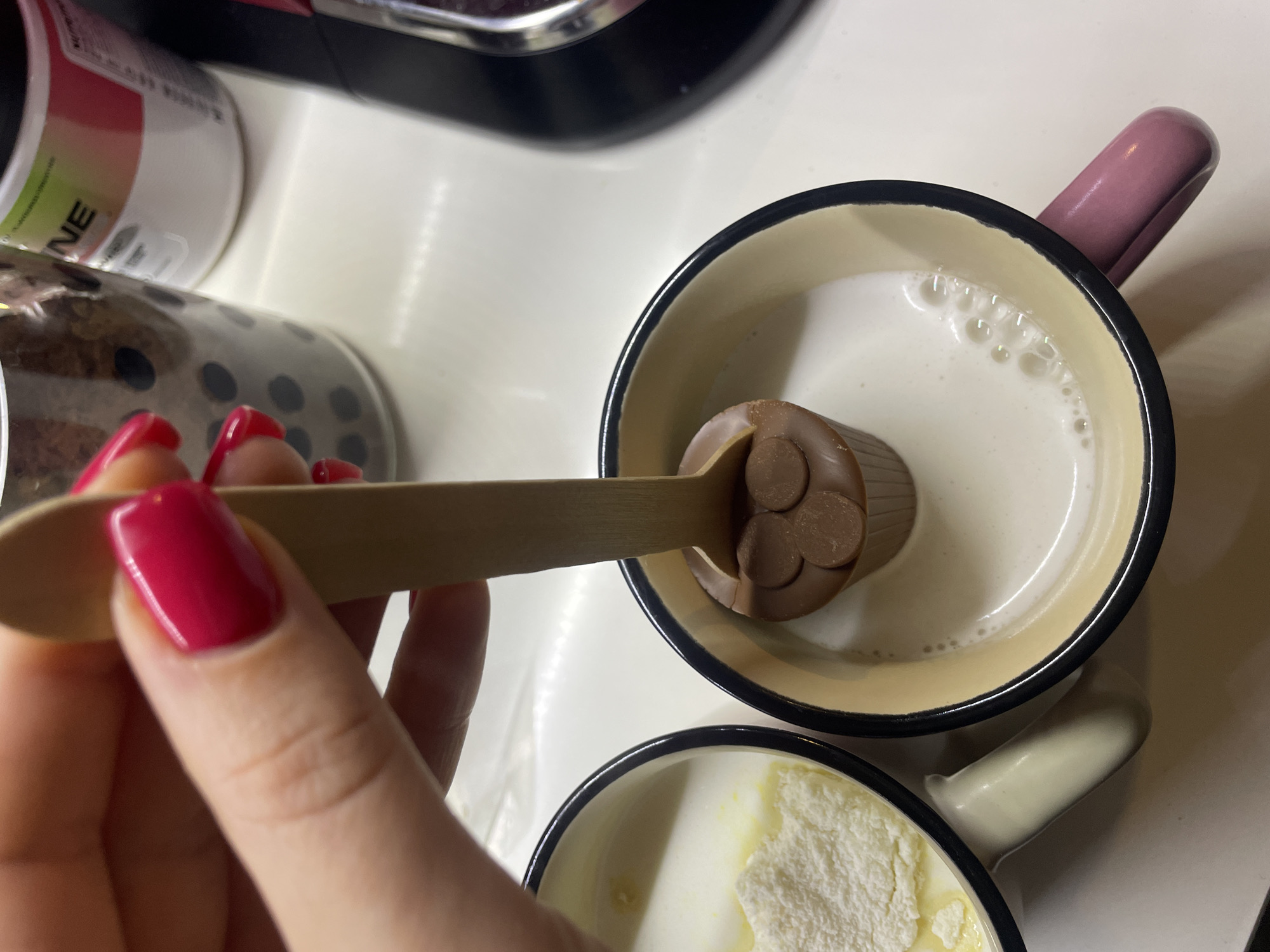 A close-up, overhead shot depicts a hand holding a wooden spoon with a chocolate stirring spoon over a mug of milk. The hand has red nail polish. Below this, another mug contains a white substance, possibly milk powder or a similar ingredient. A portion of a white container with green labeling and a jar with dark contents are visible in the background. The background includes a black and silver object as well. The scene gives no specific indication as to the location in Sofia, Bulgaria. The environment is indoors.