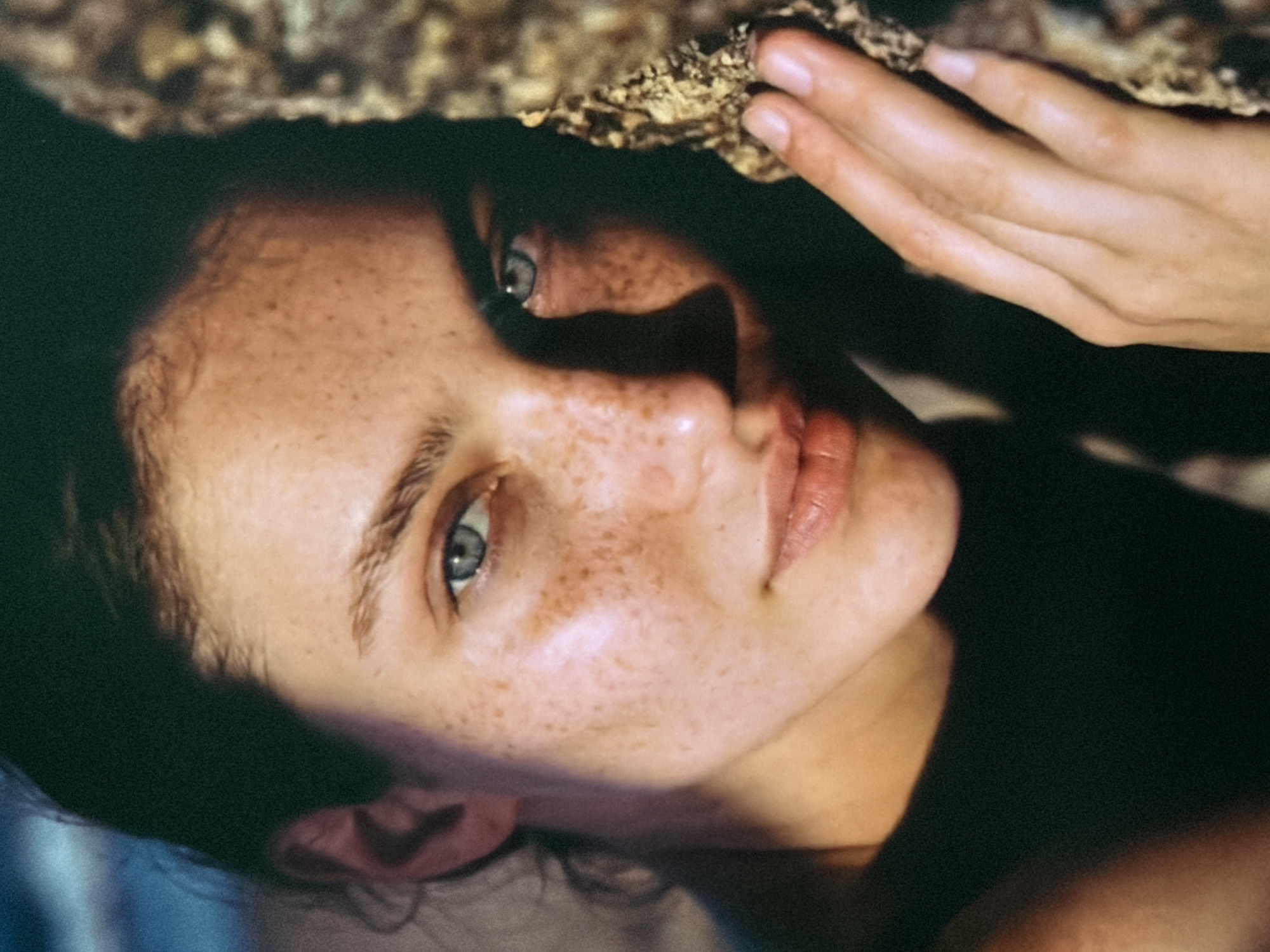 The image shows a close-up, head-and-shoulders portrait of a young woman with fair skin and numerous freckles. She has striking blue eyes and her hair is dark and pulled back. She is leaning her head against a rough, textured surface that appears to be a rock or tree bark. Her left hand is visible, resting on the textured surface near her face. The lighting creates strong shadows and highlights on her skin, emphasizing her features. The background is dark and out of focus, with a hint of blue in the upper left corner, possibly suggesting water or sky. There are no indications that this image was taken in Bobrovo, Russia.