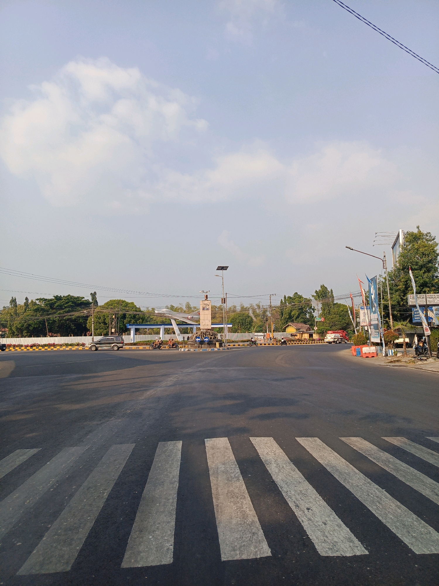 A wide, asphalt road intersection in Sugihwaras, Indonesia, features a pedestrian crosswalk in the foreground. The road is mostly empty, with a few vehicles including a silver SUV, motorcycles, and a red truck visible in the middle ground. On the left side of the intersection, a white fence runs along the edge of the road, bordering trees and buildings. A prominent landmark in the middle distance appears to be a monument with an airplane sculpture and a small building with a blue roof. Various flags and billboards are scattered throughout the scene, suggesting a commercial or public area. The sky above is a pale blue with scattered clouds, indicating daytime. The overall impression is of a typical urban intersection in Indonesia.