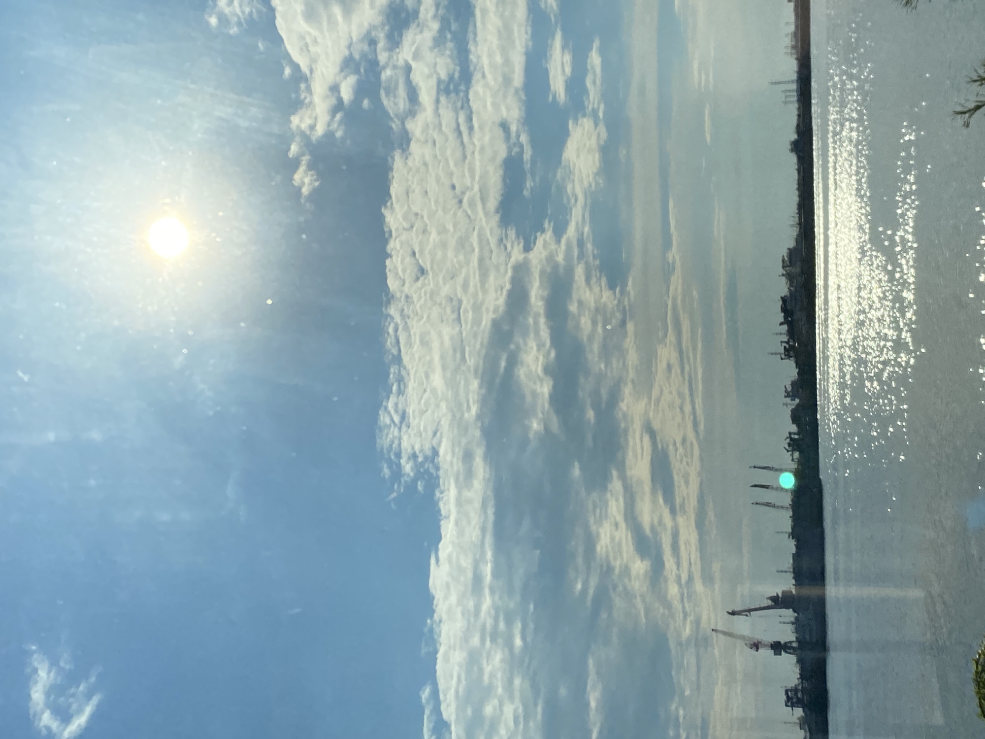 This image depicts a wide body of water reflecting the bright sun and a cloudy blue sky. Along the far shore, a silhouetted industrial landscape with numerous cranes is visible. The water's surface is characterized by numerous bright, sparkling reflections of the sun. There are no people or animals visible in the frame. The scene is consistent with an industrial port or harbor area. While the image strongly suggests an industrial port, it does not provide specific information that allows for definitive placement within Chaikovsky, Russia, or any other specific location.