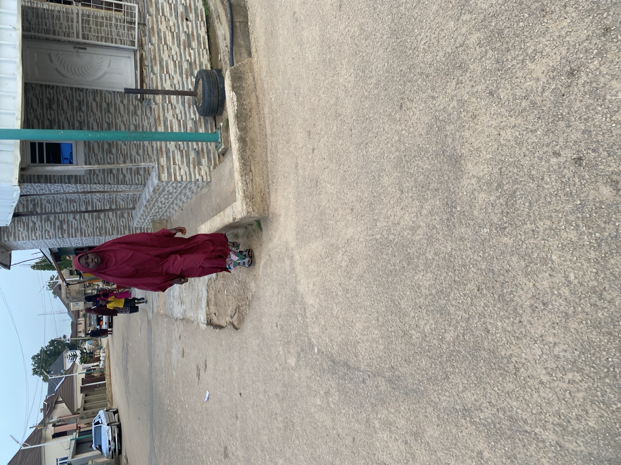 A young woman wearing a maroon hijab and dress walks down a paved street in Jos, Nigeria. She is wearing sandals. Behind her, a few other people, including a child in a yellow shirt, walk in the same direction. Cars are parked on the side of the road, and houses line the street. The sky is overcast.