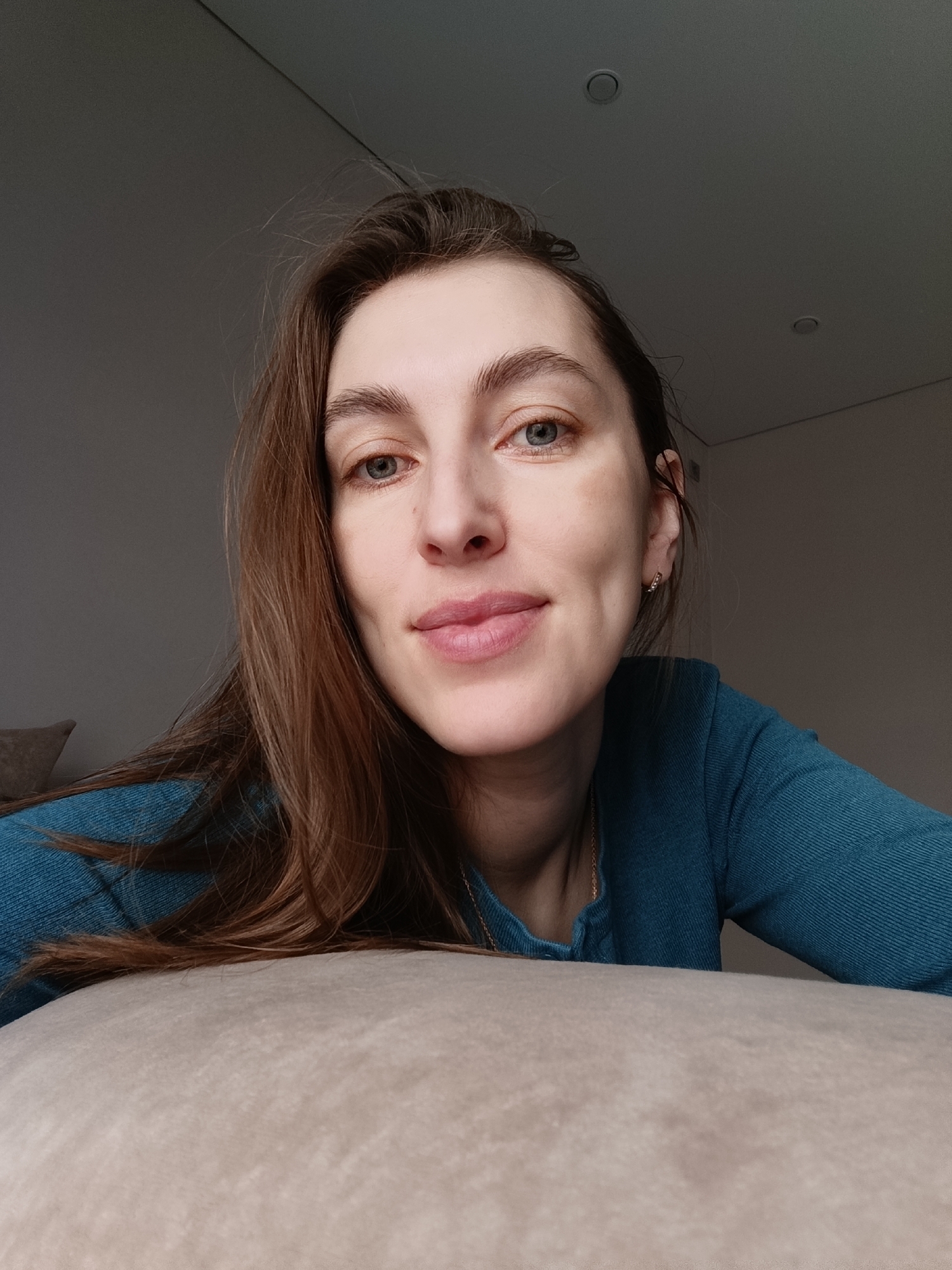 A headshot-style portrait features a young woman with long, medium-brown hair, seen from the chest up. She has fair skin, light-colored eyes (blue-grey), and a subtle, natural make-up look, including filled-in eyebrows and pinkish lips. A faint smile plays on her face, and her expression is calm and direct as she looks towards the viewer. She is wearing a dark teal or blue-green long-sleeved top, possibly made of ribbed fabric. A delicate gold chain is visible around her neck. In her right ear, a small hoop earring with what appears to be tiny stones or diamonds is visible.
The woman is positioned in the lower-middle of the frame, with her body angled slightly to the right. The foreground is dominated by a light beige or off-white cushion or pillow that she appears to be leaning on or resting against. The background consists of a plain, light grey wall on the left and a darker grey ceiling on the right side, with two recessed lights visible. The lighting is soft and natural, suggesting daytime indoors, with light appearing to come from the left, casting a subtle shadow on her right cheek and the right side of the room. The overall scene conveys a relaxed, intimate atmosphere within what appears to be a residential setting in Chaikovsky, Russia. There is no visible text in the image.